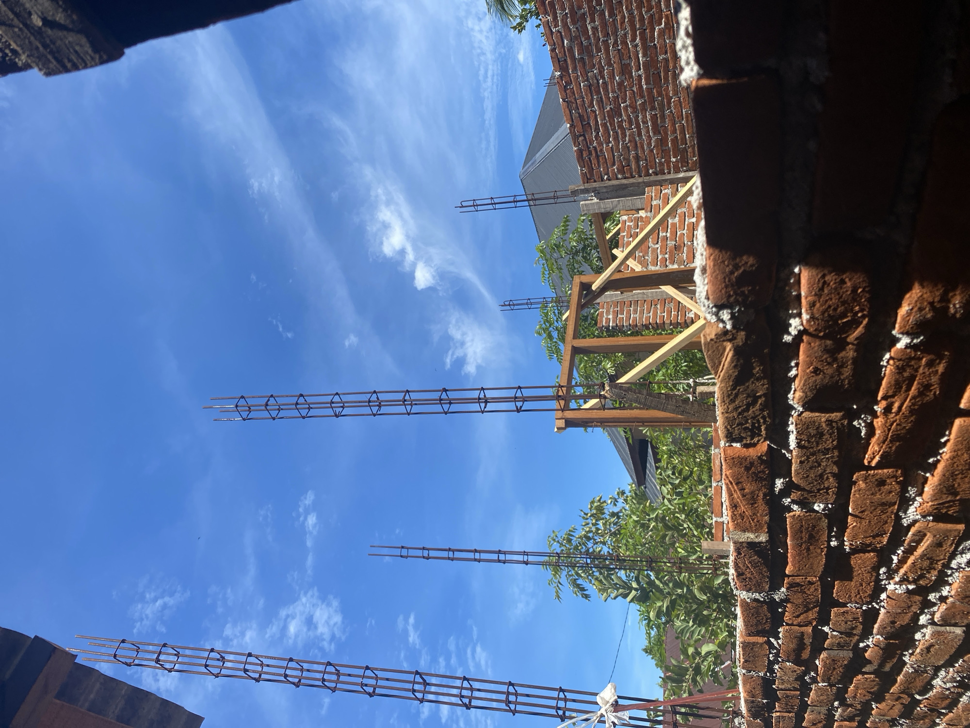 The image depicts an under-construction building in Banda Aceh, Indonesia. The foreground is dominated by a rough brick wall. Behind this wall, several vertical rebar columns rise towards a bright blue sky with scattered wispy clouds. Further in the background, more brickwork is visible, along with wooden framing elements and the dark sloped roof of another building. Green foliage from trees and plants surrounds the construction site. The scene suggests a typical day of construction activity under clear weather conditions.