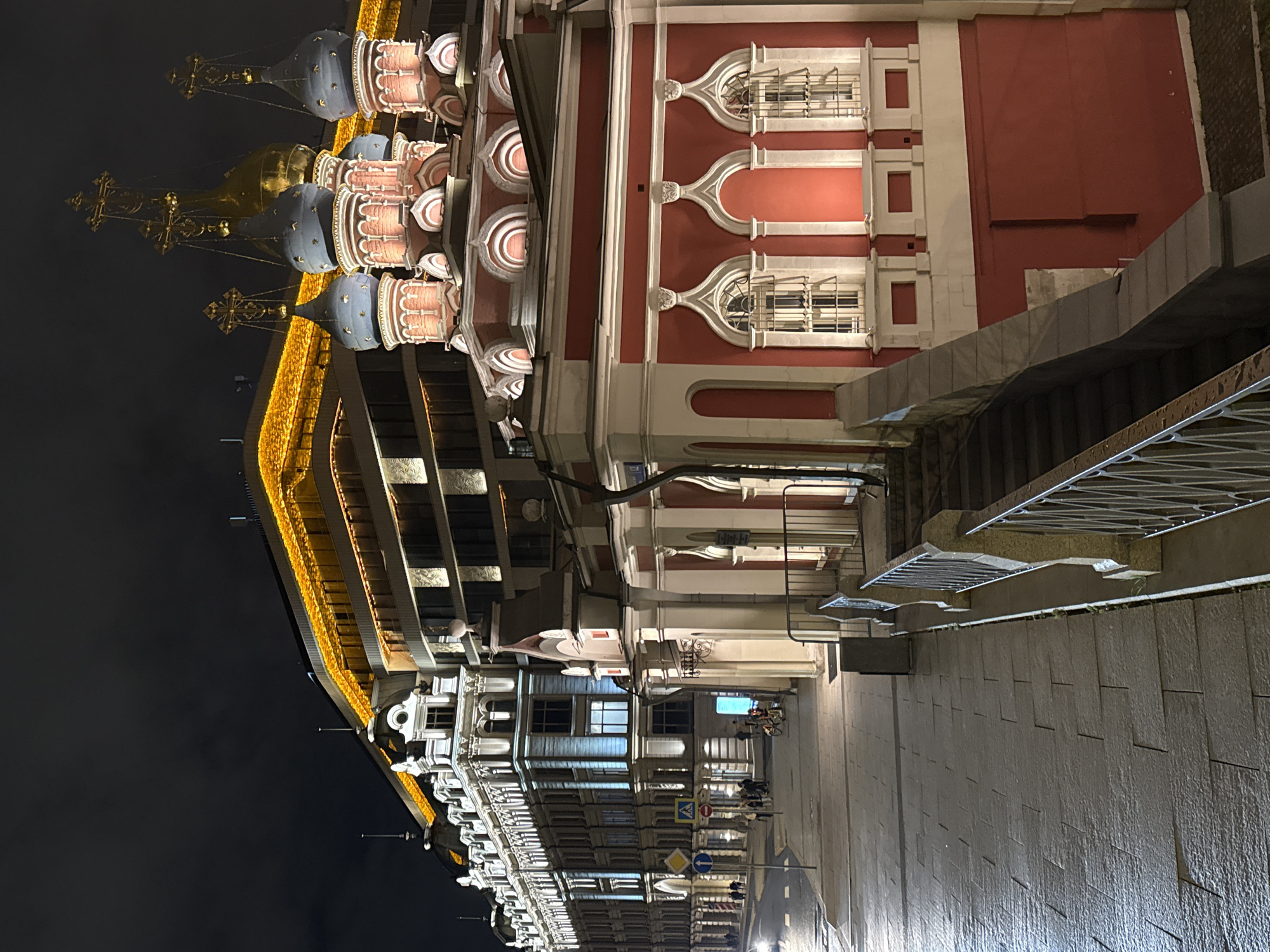 The image depicts a street scene in Moscow, Russia, at night. Illuminated buildings line the street, featuring ornate architecture and red and white facades. A distinctive structure with multiple domes and golden crosses suggests the presence of a Russian Orthodox church or cathedral. The street is paved with stone, and a set of stairs leads up to one of the buildings. People are visible on the street, although they appear small and somewhat blurred. The scene is well-lit, with the buildings and streetlights casting a glow on the surroundings.