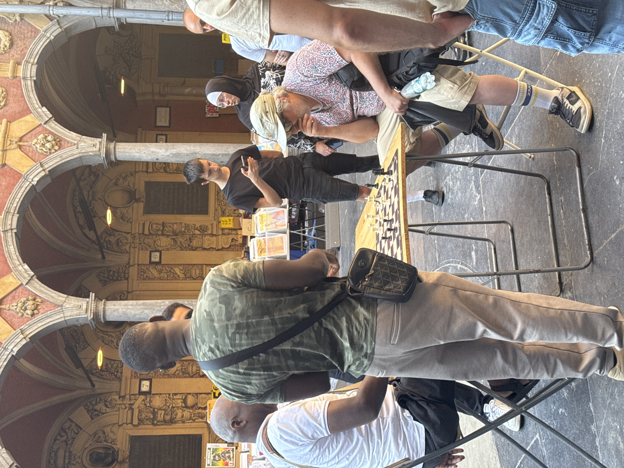 The image depicts an outdoor scene in Lille, France, possibly at an outdoor market or a public square with arcade architecture. Several individuals are present, with a primary focus on a game of chess taking place at a wooden table.
On the left, a man wearing a white t-shirt and dark pants is seated, facing away from the camera, observing the chessboard. To his right, a man in a camouflage t-shirt and light-colored pants stands, also looking towards the chessboard. He is carrying a black crossbody bag.
The center of the image features a man wearing a light-colored baseball cap, sunglasses, and a patterned t-shirt, seated and deeply engrossed in the chess game, with his hand near his chin. He is wearing khaki shorts and striped socks with sneakers. Beside him, a woman in a black hijab and glasses is standing, looking at the chessboard. To the right of the woman, a man in a white t-shirt and denim shorts is partially visible, with his hands in his pockets. Another person, only their legs are visible from the waist down, wearing denim shorts and standing on the far right of the frame.
The chessboard is set up on a wooden table, with chess pieces arranged for play. The background reveals architectural elements characteristic of Lille, including arched walkways with decorative stonework and classical columns. Several stands with books and other items are visible in the mid-ground. The ground is paved with dark stone.