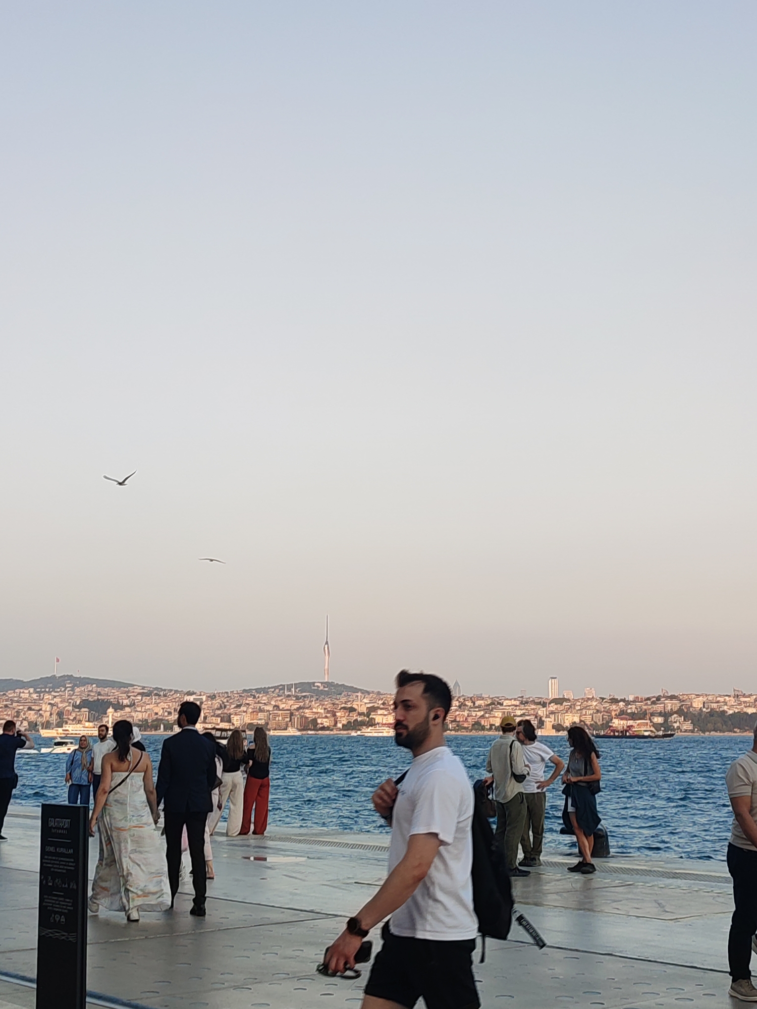 The image depicts a waterfront promenade in Beyoğlu, Turkey, with the Bosphorus strait and the city skyline in the background. Several people are present, some walking, others conversing. In the foreground, a man in a white t-shirt and black shorts walks away from the camera, carrying a black backpack. To his left, a couple is walking away, the woman in a long, patterned dress and the man in a dark suit. Further back, a group of individuals stands near the water's edge, some looking out at the strait. The water is a vibrant blue, with boats visible in the distance. The skyline is characterized by numerous buildings and a prominent tower, indicative of a major urban center. Two birds are in flight in the sky above the cityscape. The promenade itself appears to be made of concrete with a textured surface, and there is a signpost to the left. The overall scene suggests a leisurely day in a popular public space.