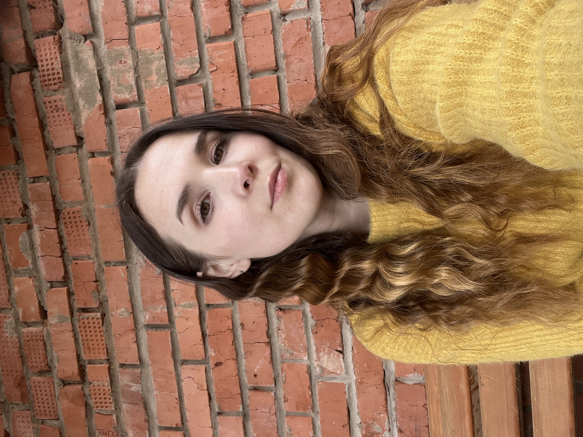 A woman with long, wavy brown hair is wearing a yellow sweater. She is positioned in front of a red brick wall, potentially an exterior wall of a building. There is a wooden structure to her right, possibly a bench.
Without additional context, it's impossible to definitively place the scene in Votkinsk, Russia. More information is required to make that determination.