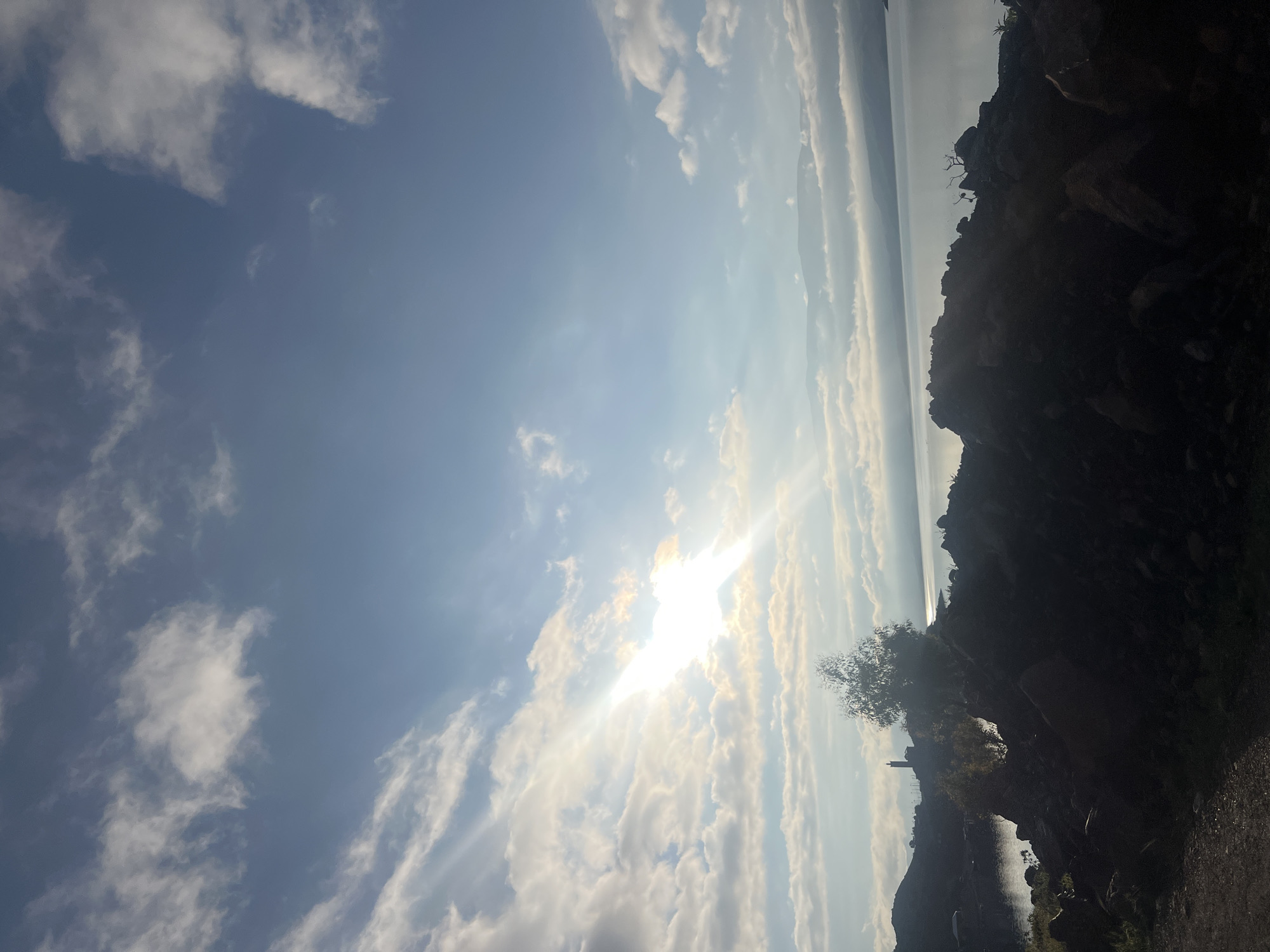 This image depicts a panoramic landscape scene at Bin El Ouidane, Morocco, likely during sunrise or sunset, overlooking the Bin El Ouidane Lake/Dam. The sky occupies the upper two-thirds of the frame, featuring a clear light blue color with scattered white, cumulus-like clouds. A bright sun is positioned in the upper-left quadrant, emitting strong rays and lens flare, illuminating the clouds from behind and casting a radiant glow.
The foreground consists of a dark, silhouetted rocky terrain, sloping slightly upwards from left to right. A dirt path is visible along the bottom-left edge. In the midground, a small, leafy tree, possibly a eucalyptus, stands prominently in silhouette against the bright light, approximately centered horizontally but in the lower-mid section vertically. To the left of the tree, on the distant shore of the lake, several structures are vaguely visible, including a tall, slender tower or minaret-like building.
The Bin El Ouidane Lake/Dam spans the middle ground and background, appearing calm and reflecting the soft light from the sky. Distant mountains or hills form the horizon line beyond the lake, partially obscured by a layer of low-lying clouds or atmospheric haze that extends above the water's surface. No individuals or overt actions are discernible within the frame.