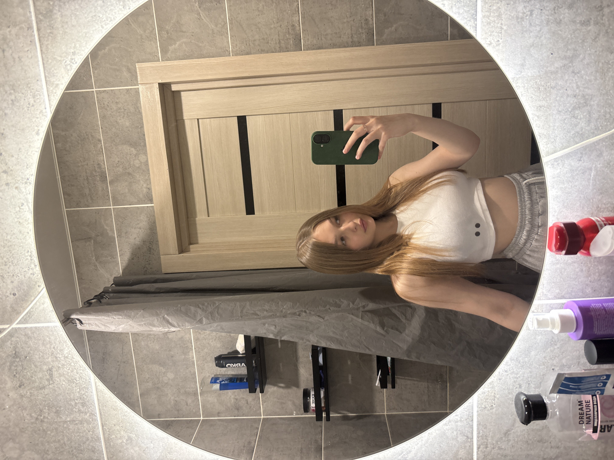 A young woman with long blonde hair is taking a mirror selfie in a bathroom. She is wearing a white crop top and grey sweatpants. Her reflection shows her holding a green phone in her right hand and wearing a ring on her left hand. In the background, there is a grey shower curtain, a wooden door, and shelves with various toiletries. The bathroom walls are tiled with grey square tiles. The image was taken in Chaikovsky, Russia.