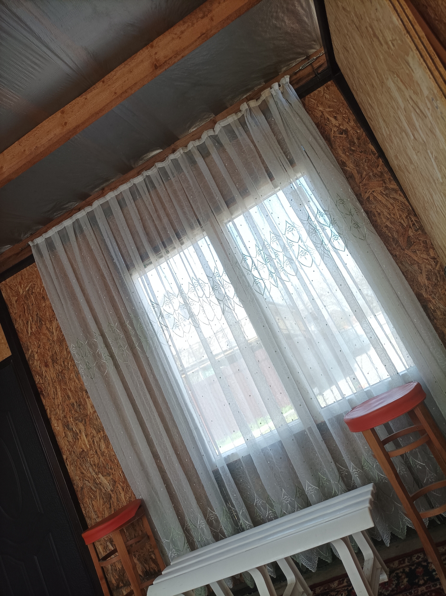 An interior shot reveals a room seemingly under construction, evidenced by exposed wood beams and unfinished walls of composite wood paneling. A window is centered in the frame, draped with sheer, patterned curtains. Two wooden stools, one with a red seat, flank a low white table or bench. The setting is likely residential, although it's not possible to confirm it's location in Ilskiy, Russia. The scene appears to be static with no evident activity taking place.