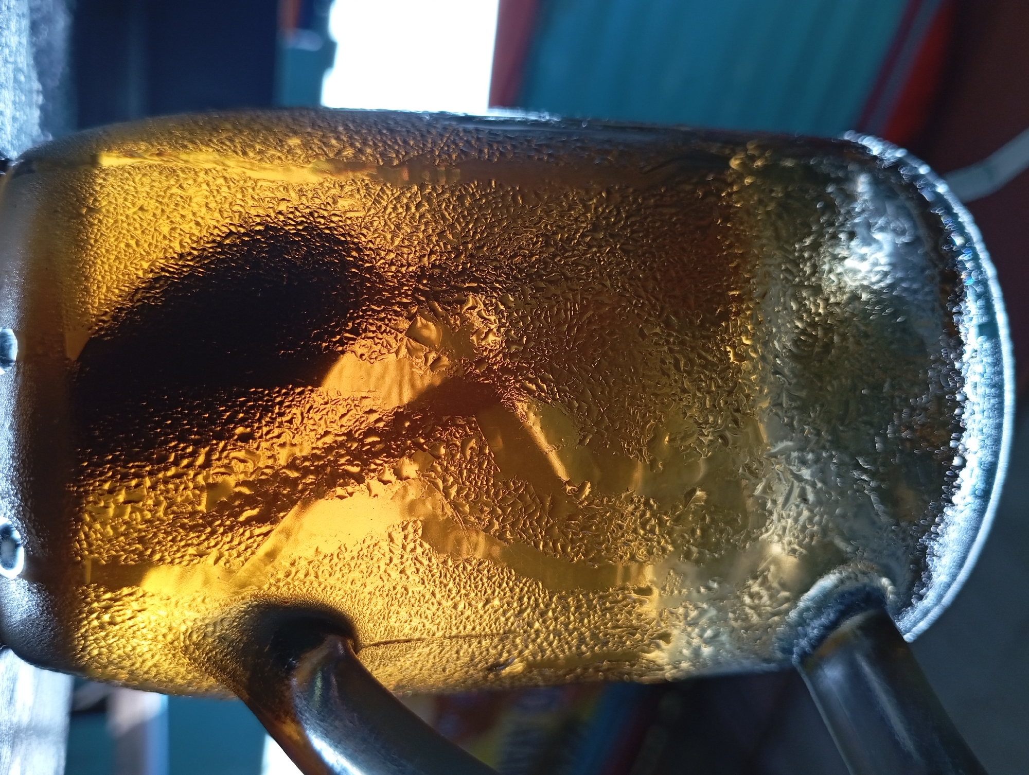 A close-up shot captures a glass mug filled with a golden-brown liquid, likely tea or iced coffee. The exterior of the mug is covered in condensation, with numerous small water droplets clinging to its surface. The light catches these droplets, creating a textured, almost crystalline appearance, especially around the rim of the mug where there's a distinct glow. A dark shadow of an object is visible inside the mug, partially obscured by the condensation and the liquid itself. The mug has a handle, also showing signs of condensation. The background is softly blurred, suggesting an outdoor or semi-outdoor setting, with hints of blue and red hues in what appears to be a casual environment, possibly a cafe or home porch. The image was taken in Lemahbang, Indonesia.