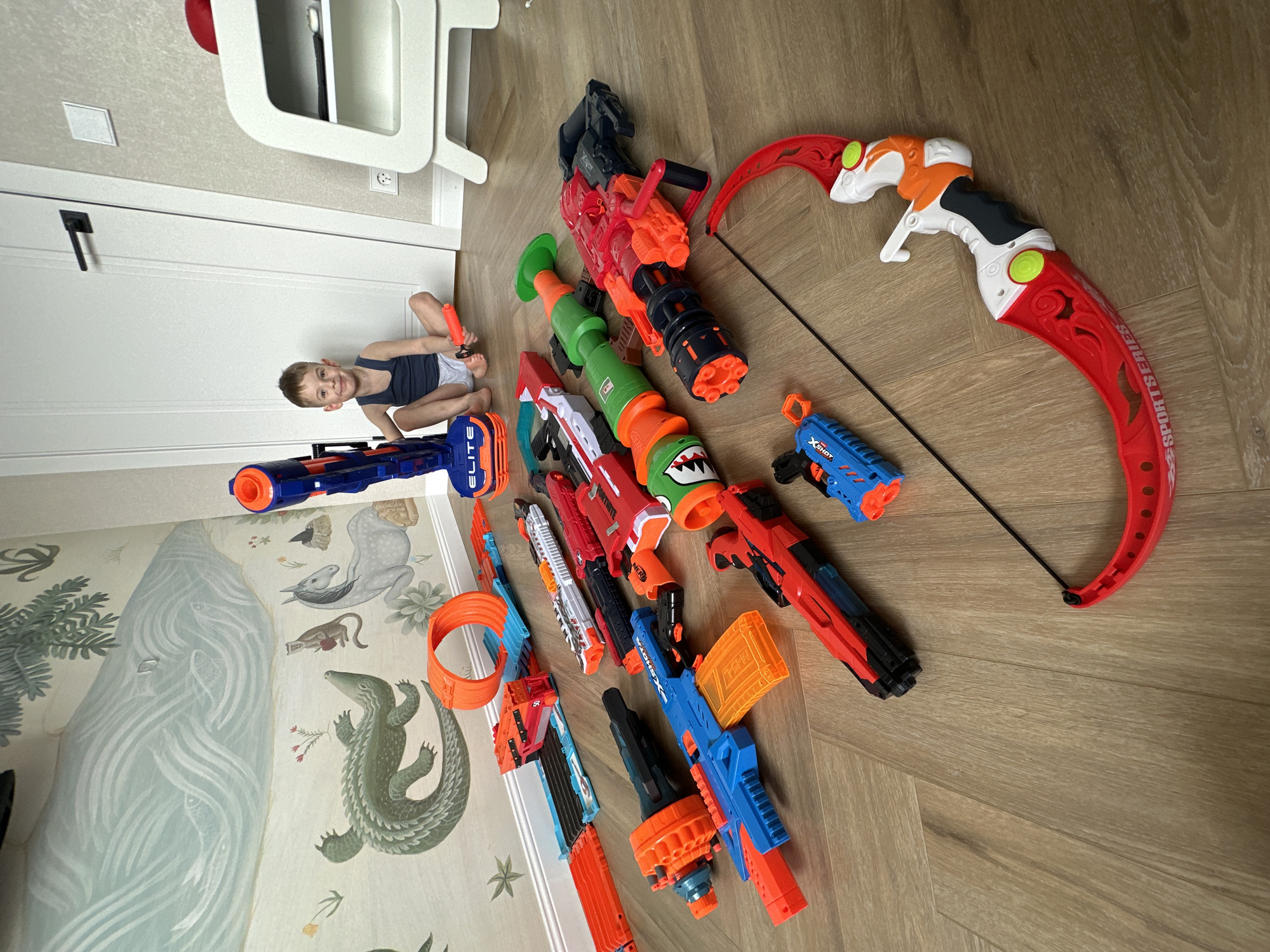A child is seated on the floor, near a white door and next to a collection of toy weapons. The toy weapons consist of multiple Nerf guns and a bow and arrow set. The floor has a light wood grain pattern. The wall to the left has a wallpaper depicting a natural scene with animals and trees. The location is Chaikovsky, Russia.