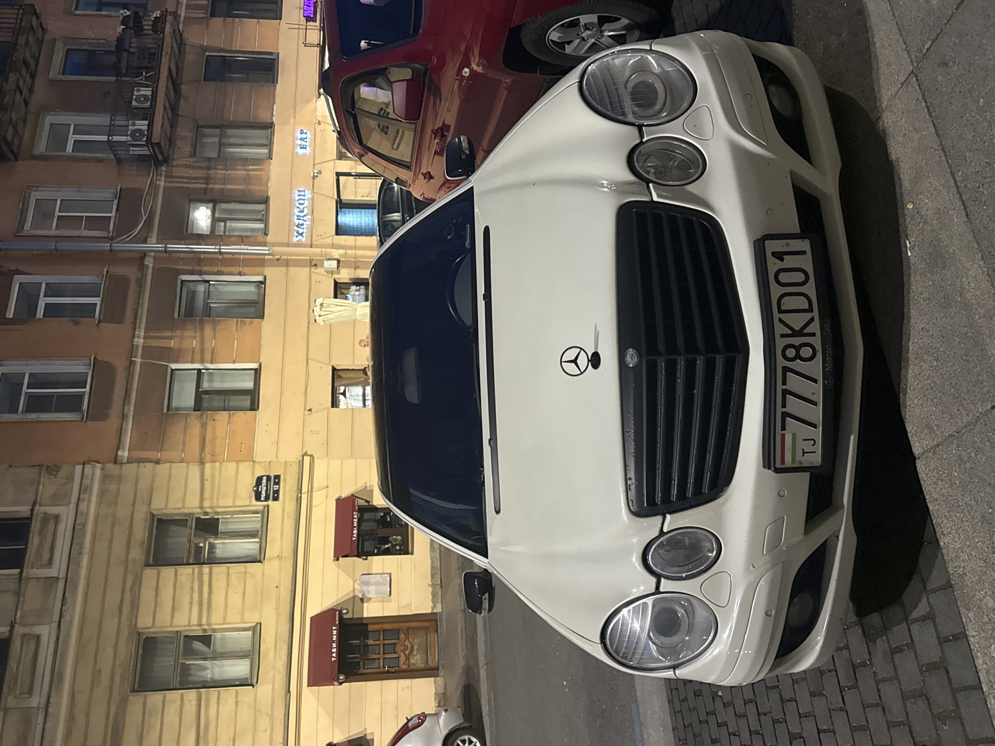 A white Mercedes-Benz E-Class is parked on a brick sidewalk in front of buildings at night. The car has a black grille and a Mercedes-Benz emblem on the hood. The license plate reads "TJ 7778KD01". To the right of the Mercedes, a red SUV is partially visible. The buildings in the background are illuminated, with windows and signs visible. One sign on a building reads "ТАБИ. МЯТ" which translates to "TAPI. MEAT", indicating a restaurant. Another sign reads "ХАЯН" in Cyrillic, which likely translates to "HAYAN" and appears to be a bar. The street appears to be cobblestone. The overall scene suggests a nighttime urban environment in Saint Petersburg, Russia.