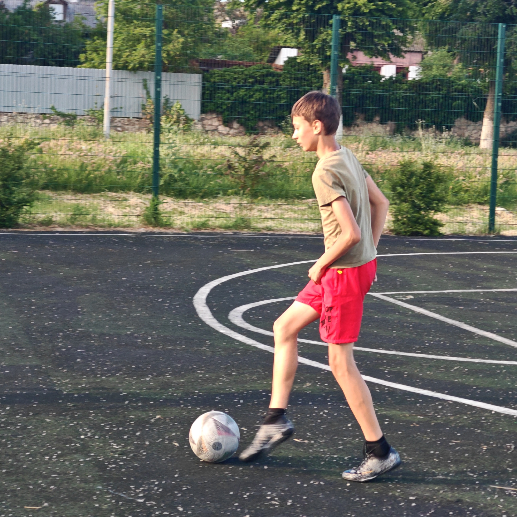 A teenage boy with brown hair wears a light green t-shirt and red shorts while playing soccer on an outdoor sports court with dark green asphalt and white lines, located in Мысовское сельское поселение, Ukraine. A white and gray soccer ball is at his feet. A green mesh fence surrounds the court, behind which is a grassy area with shrubs and trees. A house with a white fence can be seen in the background.