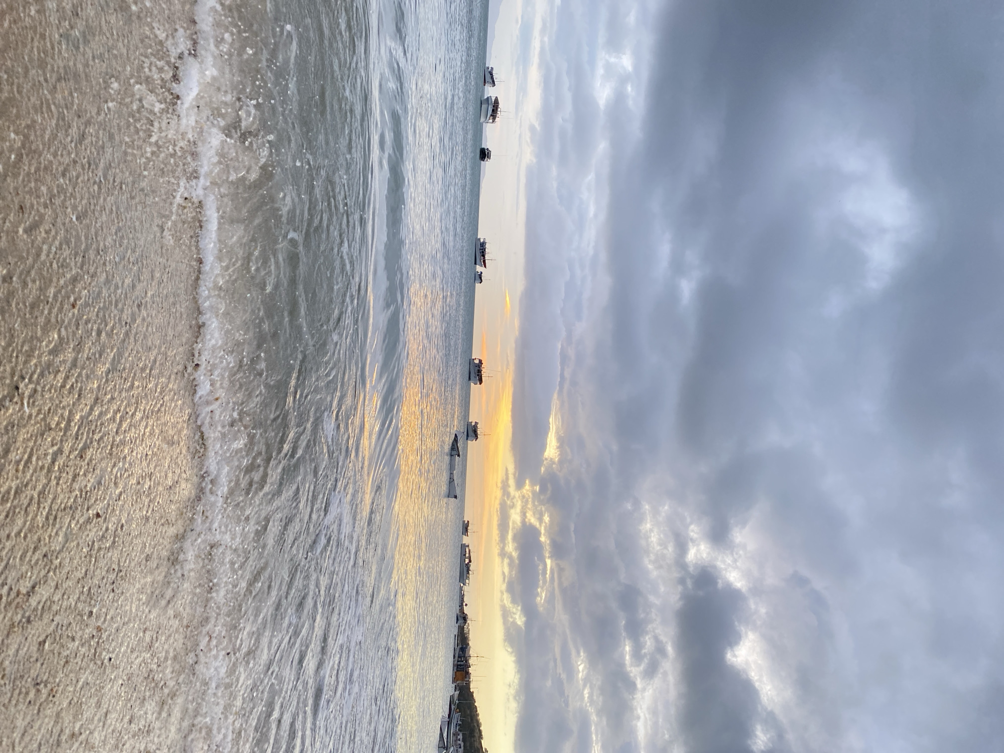 The image captures a shoreline scene in Juan Griego, Venezuela. The view is oriented vertically, with the beach on the left and the sky on the right. The shoreline displays a strip of sand meeting the water's edge, with small waves washing ashore. The water extends towards the horizon, where several boats are anchored. The sky is filled with heavy, gray clouds, with hints of a sunset glow near the horizon between the clouds and the water. The scene is still and quiet, with no visible human activity.