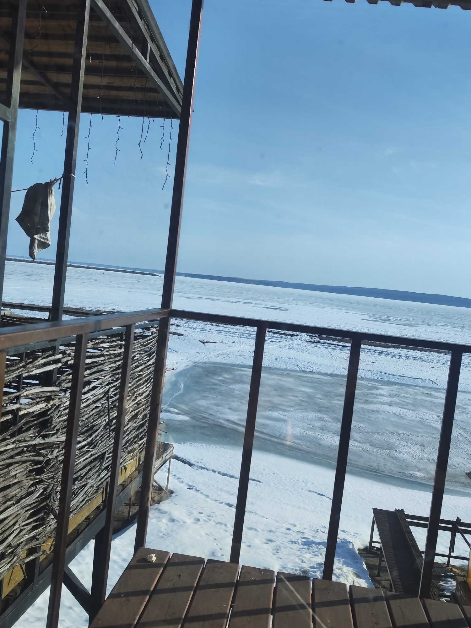 The image depicts a winter scene from a structure with a wooden deck and metal railings. The view overlooks a large expanse of ice and snow, likely a frozen body of water. In the background, a distant shoreline is visible against a clear blue sky. The location is Chaikovsky, Russia. A garment hangs from a line attached to the structure. The structure itself appears to be a deck or balcony.