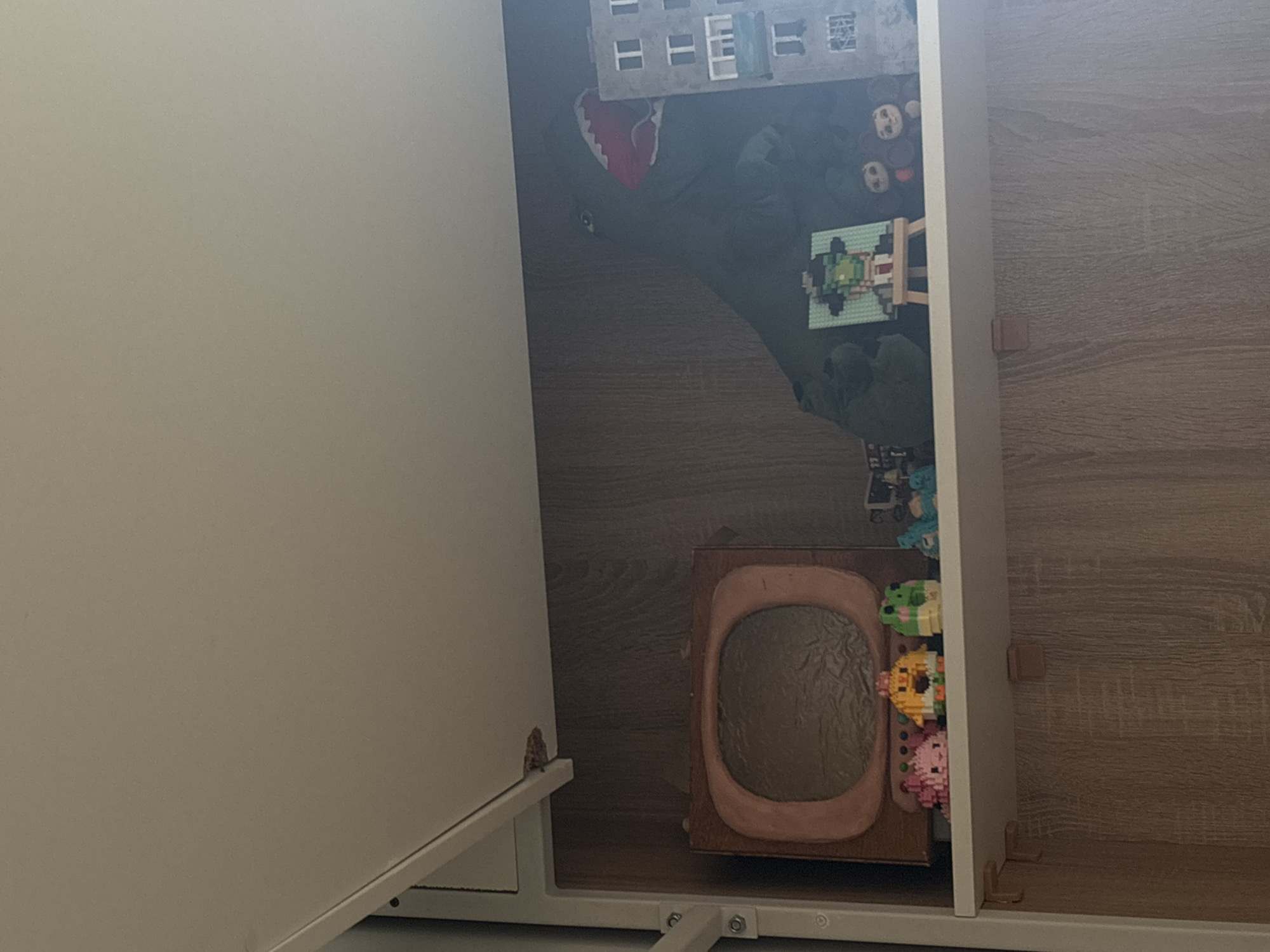 The image depicts an open cabinet in what appears to be an interior space. The cabinet's door is off-white, while the interior is a wood-grain finish. The contents of the cabinet include toys, such as a building set, a collection of small pixelated animal figures, and a framed brown object with a dark center. Other unidentified items, possibly clothing or soft materials, are also visible. The location in the city of Perm, Russia, cannot be determined from the given image.