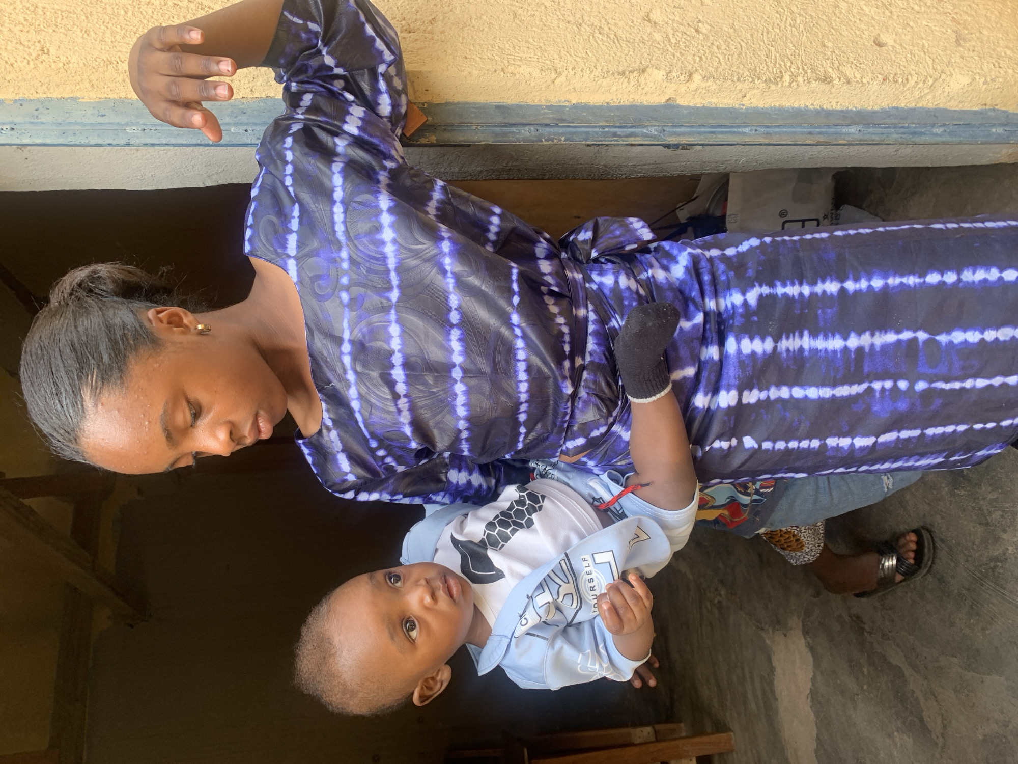 In Gura Top, Nigeria, a young woman stands in a doorway holding a baby. The woman is wearing a blue and purple tie-dye dress. The baby is wearing a light blue outfit with a white and black graphic on it. The background shows a dark interior space and a yellow exterior wall.