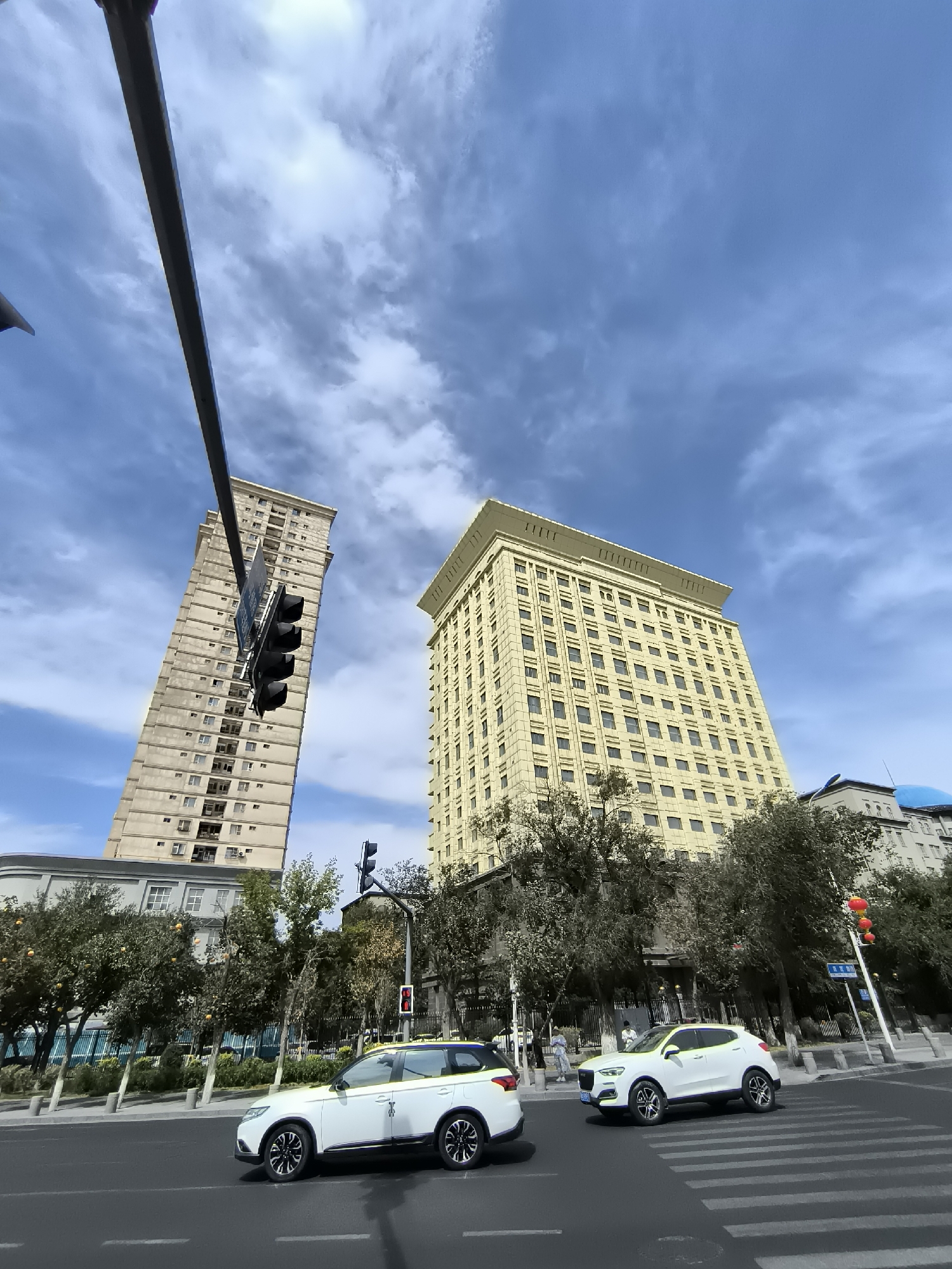The image captures a street scene in Ürümqi, China, on a partly cloudy day. Two multi-story buildings dominate the background. The building on the left is a tall, slender, beige apartment building with numerous windows. To its right is a wider, cream-colored building with a more ornate facade, also featuring many windows. In the foreground, a street with a crosswalk is visible. Two white SUVs are driving on the road. The SUV on the left is partially visible, while the SUV on the right is crossing the pedestrian walkway. Several trees with some orange fruits are planted along the sidewalk. A traffic light with red and possibly other signals is visible. The sky is a clear blue with scattered white clouds.