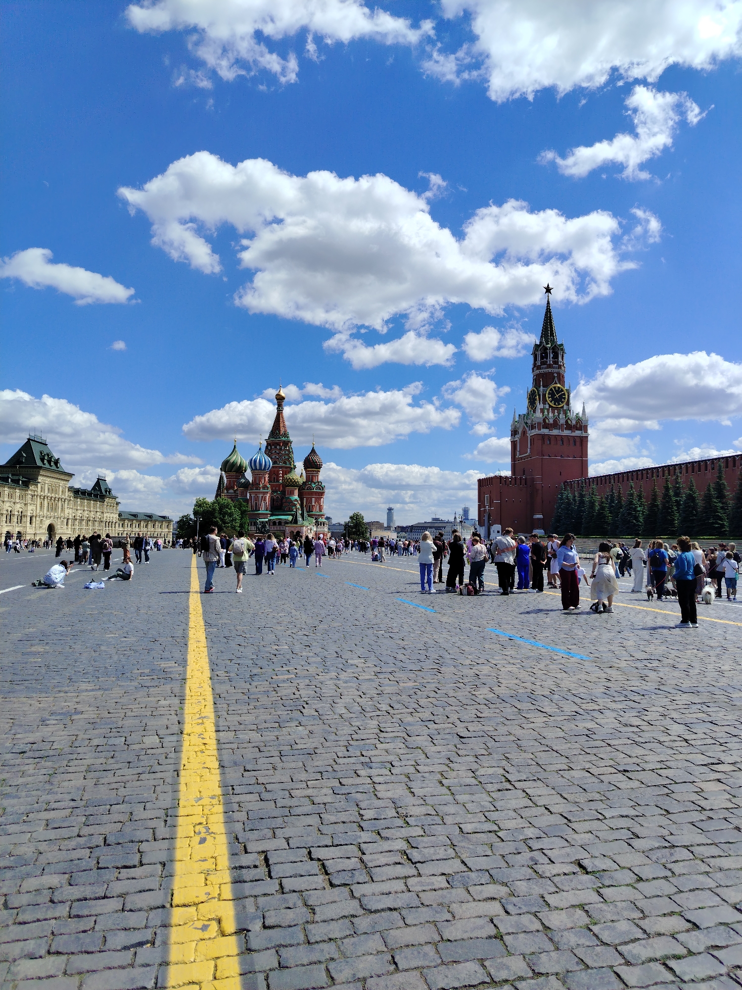 The image shows a wide cobblestone plaza under a bright blue sky with scattered white clouds. In the background, several iconic buildings are visible: a large, ornate building on the left, St. Basil's Cathedral with its colorful onion domes in the center, and a tall red brick clock tower on the right, part of the Kremlin wall. A line of evergreen trees runs along the top of the Kremlin wall.
Numerous people are spread across the plaza, appearing to be tourists. Some are walking, others are standing in groups, and a few are sitting or kneeling on the cobblestones. There are also some blue painted lines on the ground, possibly indicating a queue or designated area. A thick yellow line runs diagonally across the foreground of the plaza. The overall scene depicts a bustling public space on a sunny day, likely a popular tourist destination.