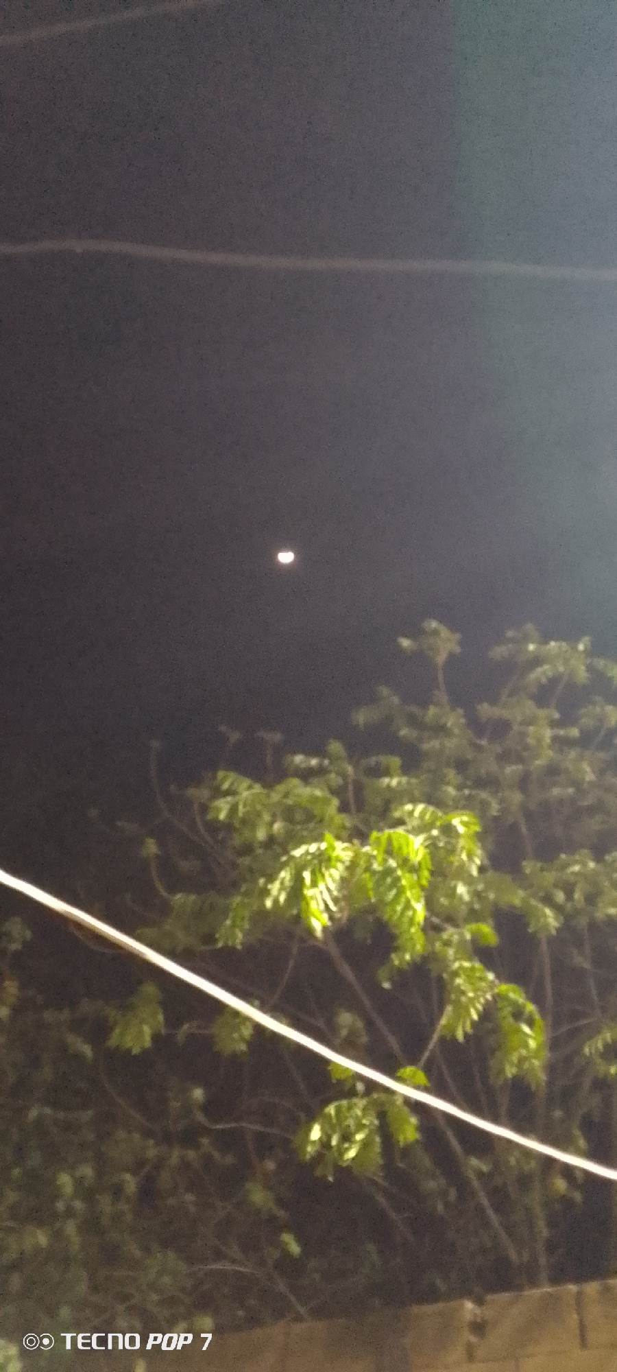 The image shows a night scene in what appears to be Altagracia, Venezuela. The sky is dark with a partial moon visible. Trees with green foliage are in the foreground. An electrical wire runs across the frame. The lower left corner contains the logo of a "TECNO POP 7" phone.