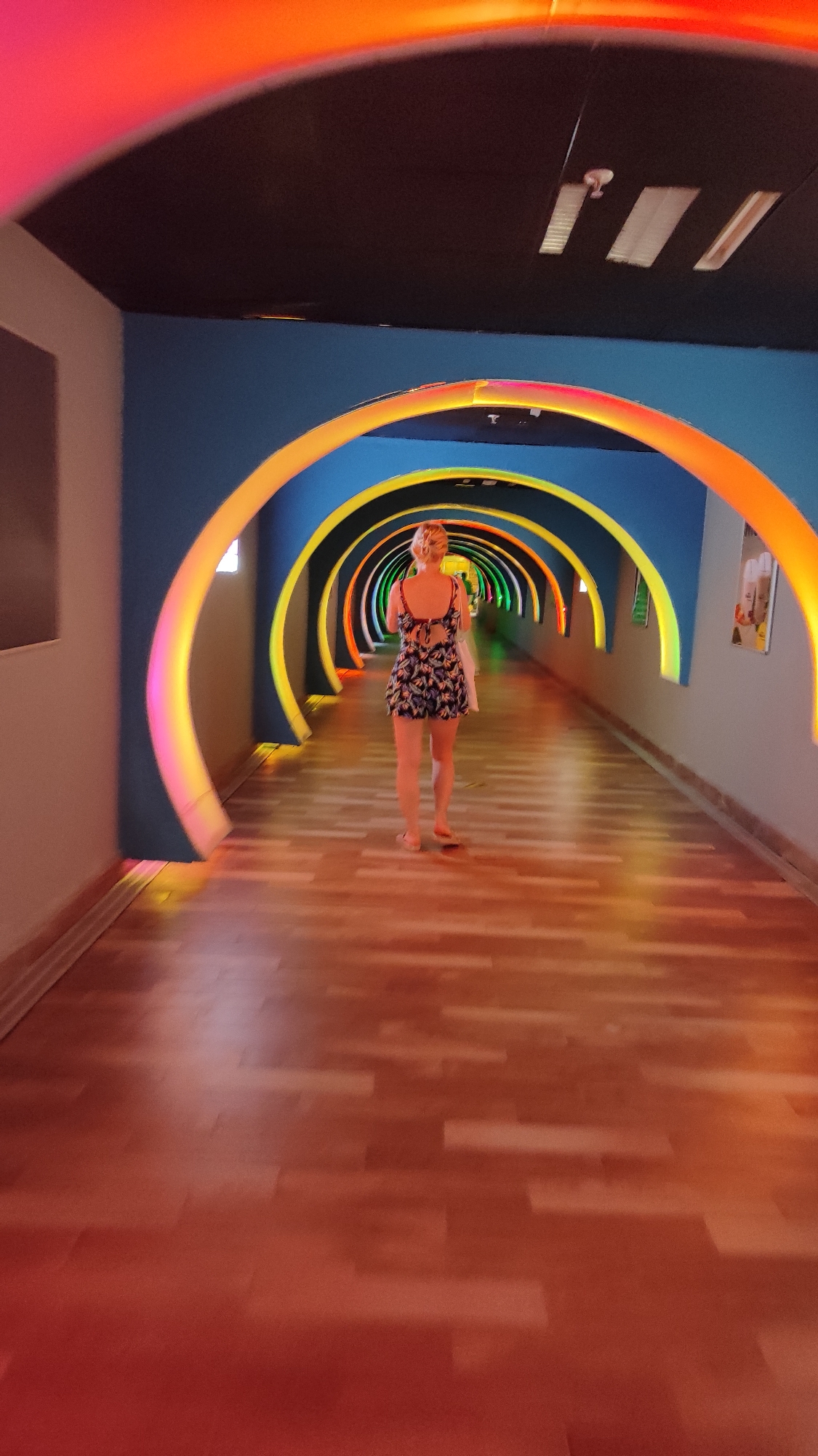 A woman is walking down a brightly lit corridor. The walls of the corridor are blue with a series of rainbow-colored arches overhead. The floor is made of wooden planks. The ceiling is dark and has three rectangular vents. The photo was taken in Alanya, Turkey.