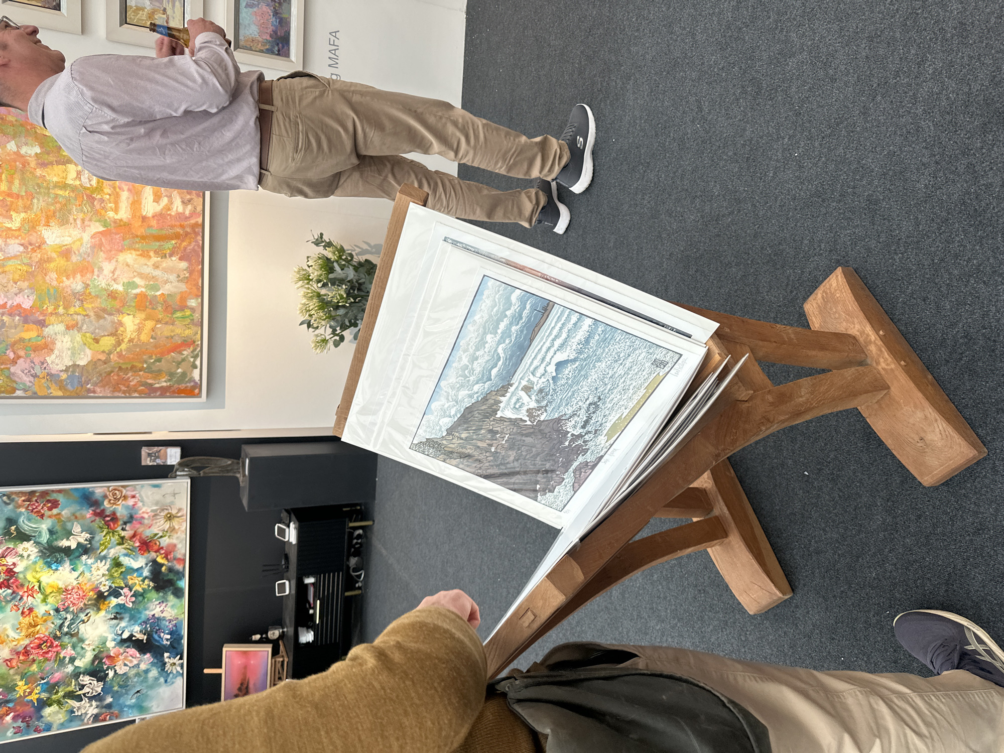 The image shows an indoor art exhibition. A man in a light purple shirt and khaki pants stands near a white wall with the name "MAFA" on it. He is looking at paintings. To the left are other abstract paintings in colorful floral patterns. In the foreground, a wooden stand displays framed artwork protected in clear plastic sleeves. A person with a tan coat and dark bag is looking at the artwork. The setting may be in London, United Kingdom, as indicated by the presence of the name MAFA, which could be a reference to the Marchand d'Art et Fabricant d'Art, an art gallery in London. The floor is covered in gray carpet.