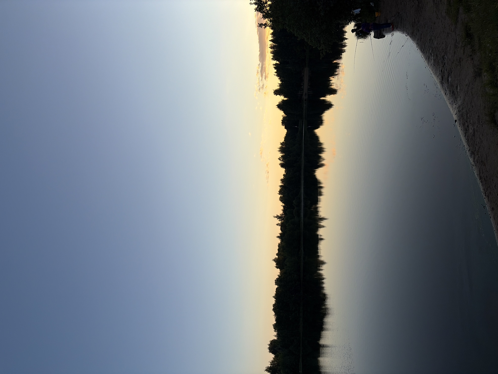 The image depicts a serene lakeside scene at dusk. The sky transitions from a clear, pale blue on the left to soft hues of yellow and pink near the horizon on the right, suggesting the setting sun. A dense line of dark trees forms a silhouette along the far bank of the lake, their reflections stretching across the still water. The water's surface is disturbed by gentle ripples, creating patterns that catch the fading light.
On the right side of the image, a sandy bank slopes down to the water. Two people are visible sitting on the bank, with fishing rods extending into the lake. One person is wearing a purple jacket and blue pants, and the other is wearing a yellow shirt. The overall mood is peaceful and tranquil, evoking a sense of quiet enjoyment of nature. The time of day is clearly evening, with the soft, diffused light characteristic of twilight. No text is visible in the image.
