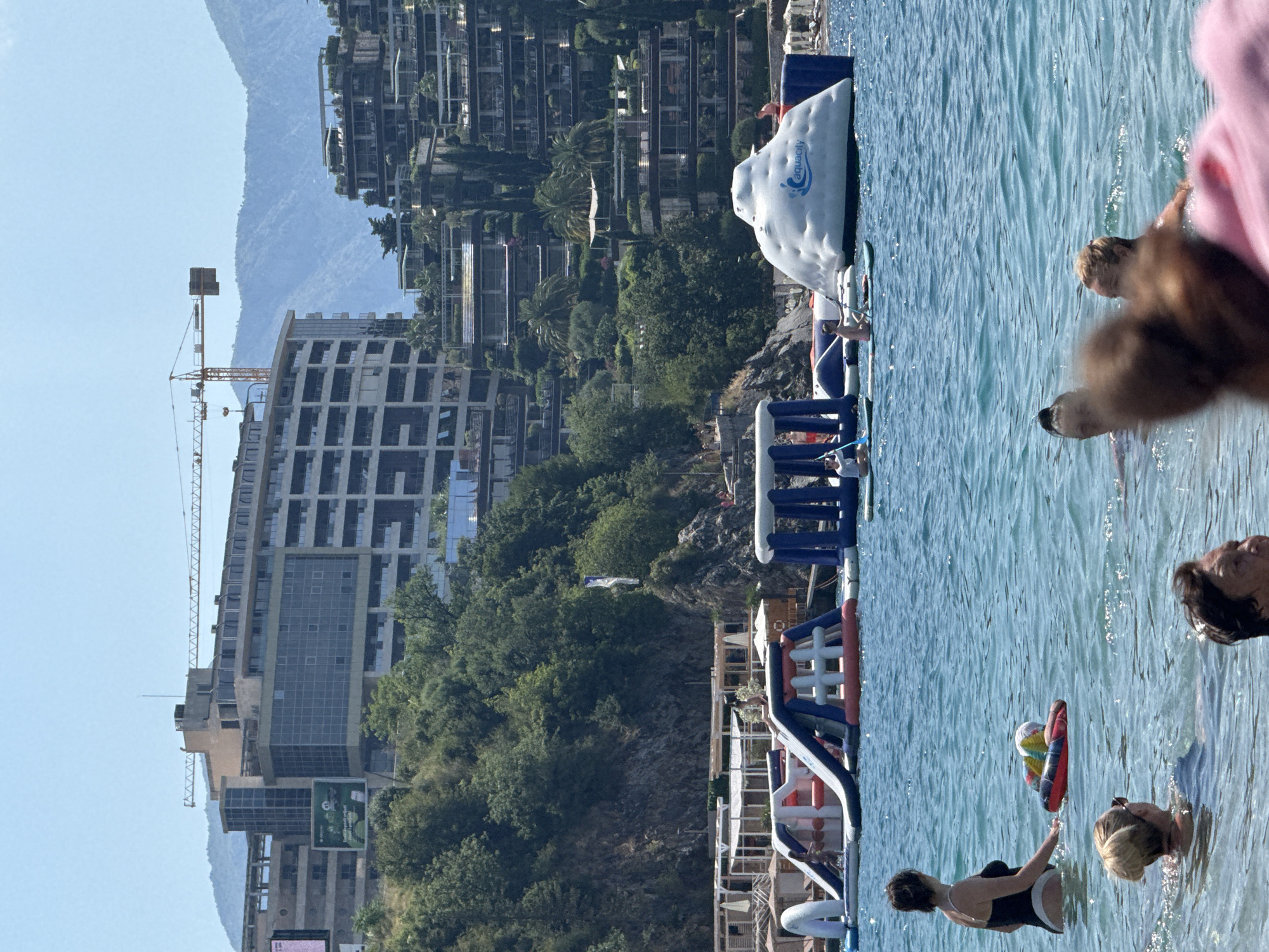 The image depicts a sun-drenched scene at a beach in Bečići, Montenegro. The turquoise water is populated with several individuals swimming and engaging in water activities. In the foreground, three people are partially submerged in the water. One woman, wearing a black swimsuit, is standing and holding a colorful float. Behind her, another woman with blonde hair pulled up is swimming, and further in the foreground, a person's head is visible, out of focus.
In the middle ground, an inflatable water park is set up. It features various structures, including slides and climbing elements, in bright blues, reds, and whites. Several people are visible on and around these inflatables, including one individual on a paddleboard. To the right of the inflatables, a large white inflatable structure with the "aquacity" logo is prominent.
The background reveals a lush, green hillside covered in trees. Behind the trees, a series of modern, terraced buildings cascade down the slope, suggesting a resort or residential complex. Further in the distance, majestic mountains rise against a clear blue sky. A construction crane is visible atop one of the larger buildings, indicating ongoing development. Beach umbrellas and structures are also present along the shoreline. The overall impression is one of a lively summer day at a popular coastal destination.