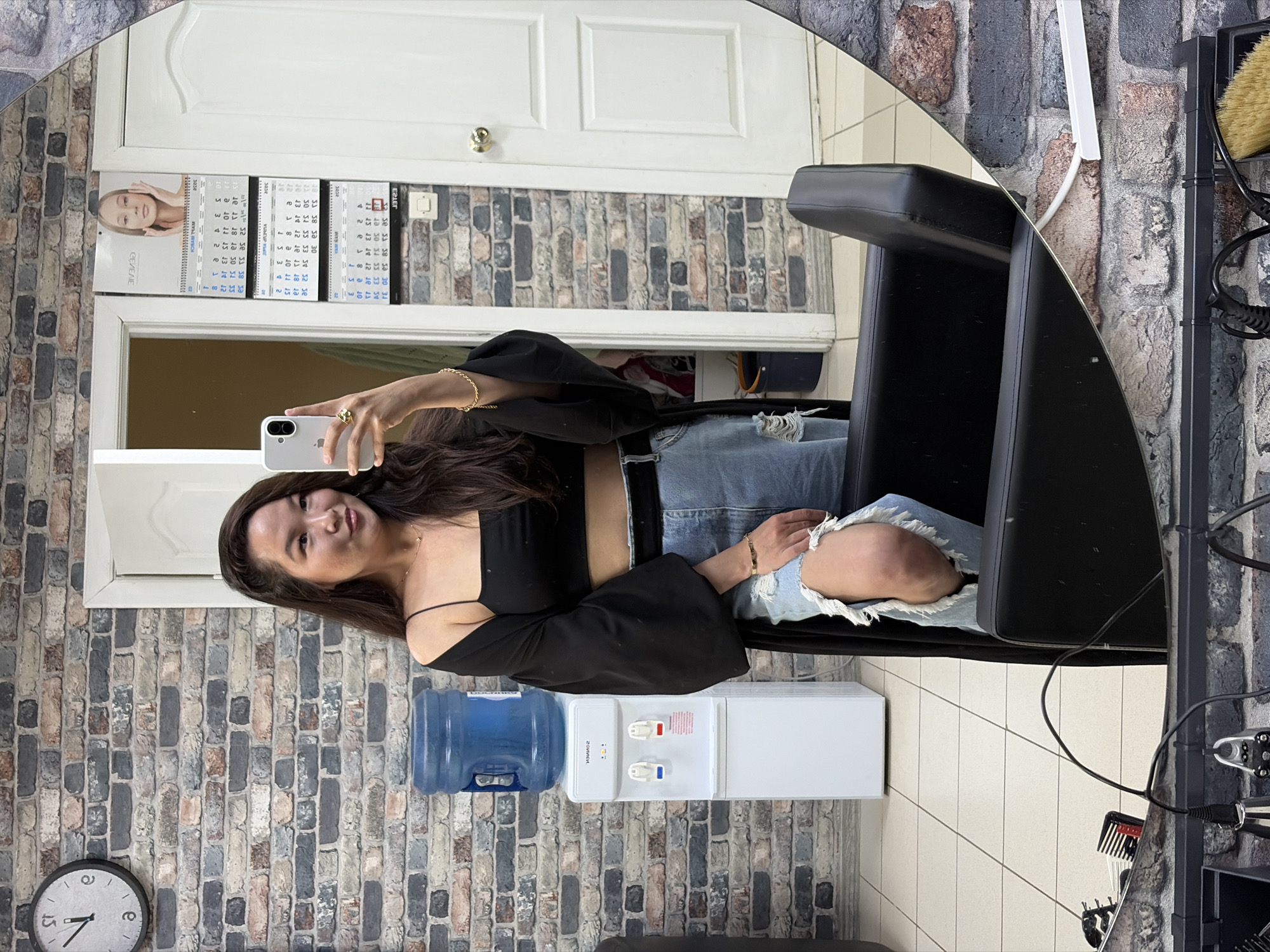 The image shows a woman reflected in a mirror. She is wearing a black top with off-the-shoulder sleeves and distressed denim jeans. She is holding a phone in her hand as if taking a selfie. Behind her, a brick wall is visible, with a clock on it. Next to the wall, there's a water cooler. The background includes a white door with a calendar hanging on it and a black salon chair. The image appears to be taken in a beauty salon or similar establishment. No specific information in the image allows the determination of the location in the city of Chaikovsky, Russia.