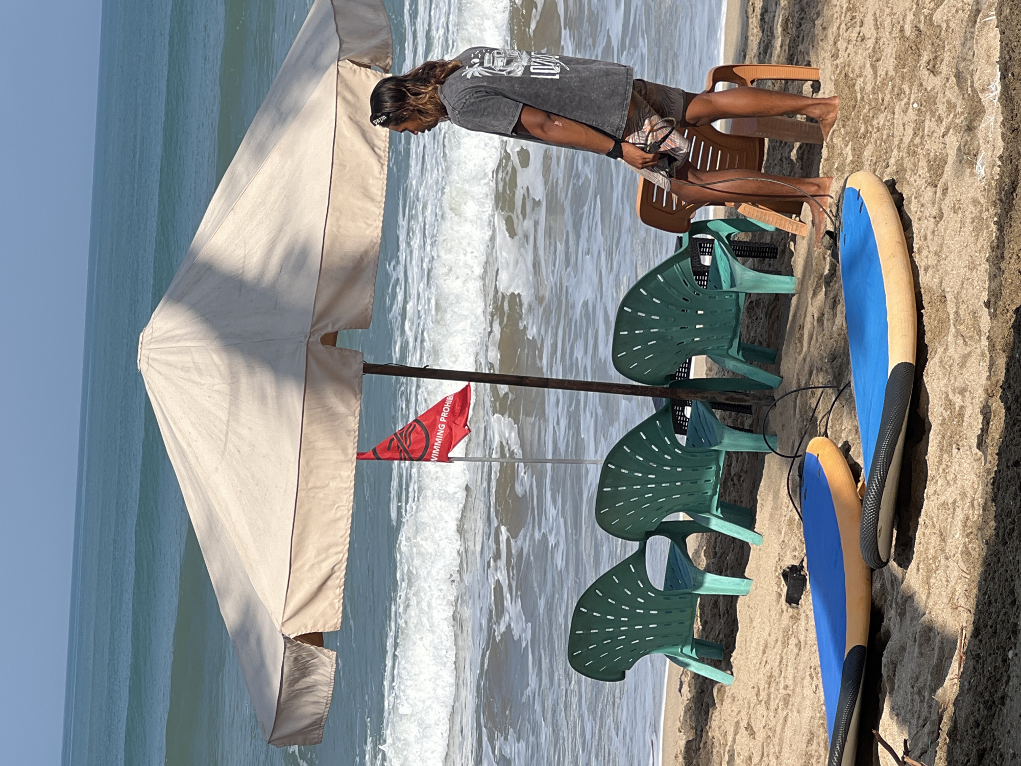 A man with long hair stands on a sandy beach in Kuta, Indonesia, under a large, light-colored umbrella. He is wearing a dark t-shirt with a graphic print and patterned shorts. He is holding a black cord in his right hand. Behind him, the ocean has moderate waves and the sky is clear. In front of the umbrella, there are three green plastic chairs and one orange plastic chair. Two surfboards, one blue and orange and the other mostly blue, lie on the sand in the foreground. A red flag with white text and symbols is planted in the sand behind the chairs.
