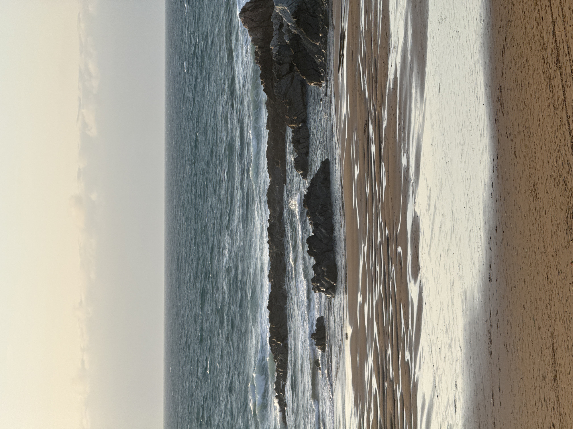 The image depicts a coastal scene at low tide on a beach in Sopela, Spain. In the foreground, the wet sand is marked with intricate patterns of shallow pools of water, reflecting the overcast sky. Further back, dark, rocky outcrops emerge from the sea, with waves gently breaking around them. The ocean stretches to the horizon under a pale, muted sky. The overall impression is one of a calm, natural landscape.