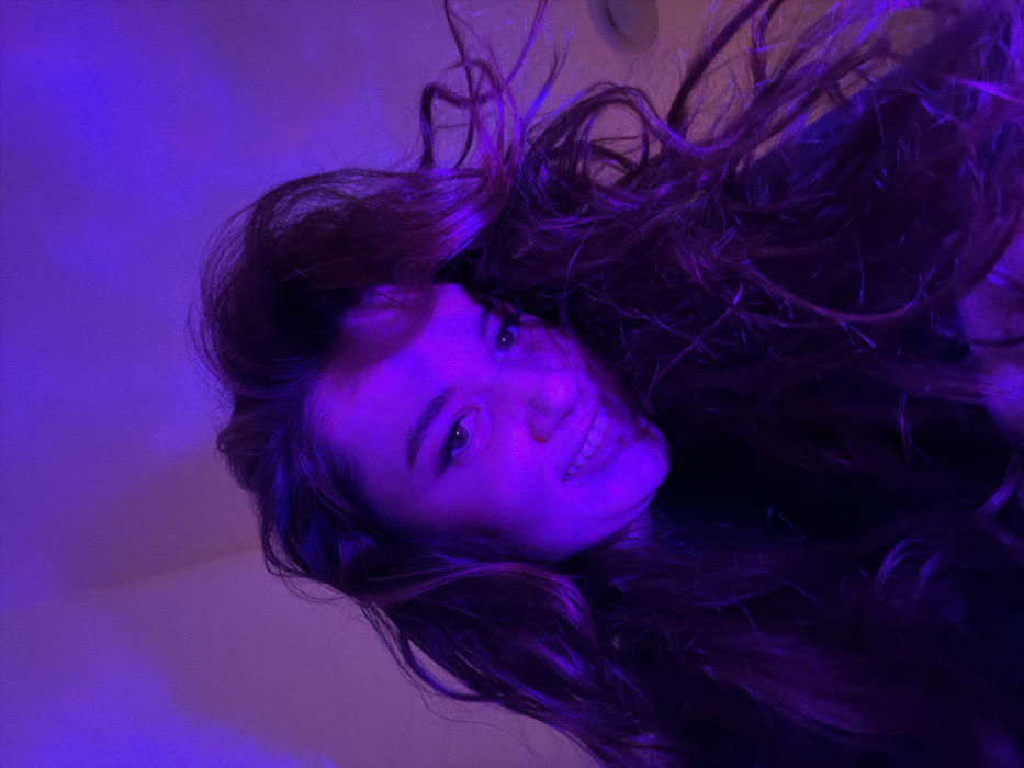 A woman with long, wavy brown hair is smiling into the camera. She is bathed in a strong purple light, obscuring some of her features. Her face is visible, including her eyes, nose, and mouth. The background is out of focus and also purple.