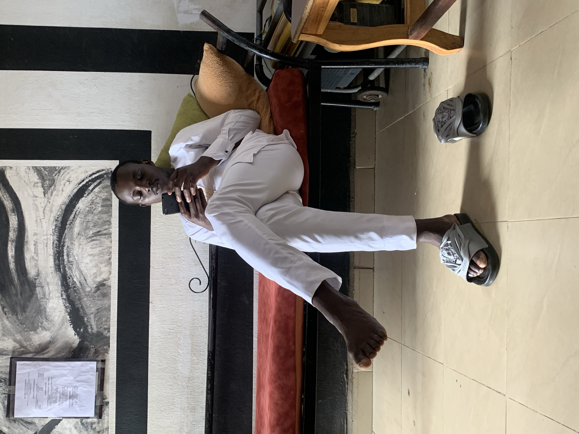 A young man, appearing to be of African descent, is seated on a bench indoors, engaged with his smartphone. He is wearing traditional white attire, consisting of a long-sleeved shirt and matching pants, paired with gray slides. The bench he sits on features a red cushion and a dark frame, with pillows behind him in muted green and orange hues. The background displays a striking mural of black and white abstract art above a black horizontal stripe. The flooring is tiled in a beige tone. To the right of the bench, a wooden table with a curved leg is partially visible. The man's posture is relaxed, with one leg crossed over the other, and his left foot is bare, while his right foot is in a slide. The overall lighting suggests an indoor environment with natural light filtering in. The context of Jos, Nigeria, is not evident from the visual information within the image itself.