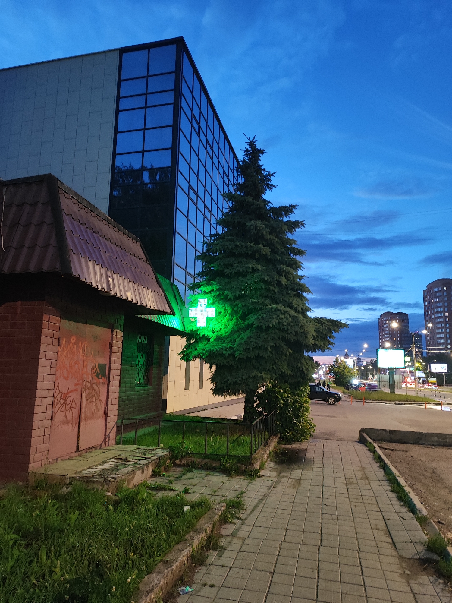 The image depicts an outdoor scene at dusk in an urban environment. In the foreground, a small, brick building with a corrugated metal roof stands to the left, featuring graffiti on its large, weathered doors and a barred window. Adjacent to this structure, a tall evergreen tree is positioned prominently, its branches illuminated by a bright green, glowing cross sign, indicating a pharmacy.
Behind the evergreen tree and the brick building, a modern, multi-story building with a facade of pale grey panels and large, dark-tinted glass windows dominates the background. The sky above is a deep blue, characteristic of twilight, with scattered clouds.
To the right of the scene, a paved sidewalk stretches into the distance, leading towards a street with visible traffic and streetlights. Several vehicles, including a dark SUV, are present on the street. Further in the background, taller residential buildings are visible. The ground alongside the sidewalk is covered with grass and some debris. The overall impression is that of a commercial area at the end of the day.