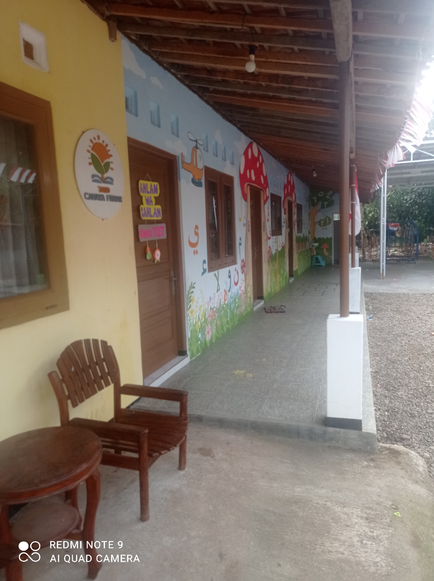 The image depicts an outdoor walkway in front of a yellow building, likely a school or community center, in Cipawon, Indonesia. The walkway is covered by a wooden-slatted roof supported by brown wooden posts. The walls of the building are adorned with colorful murals featuring children's drawings of clouds, mushrooms, a helicopter, trees, and Arabic calligraphy. Several doors line the wall, each with a different sign.
On the left side of the walkway, a wooden chair and a round wooden table are placed on a concrete floor. The chair is made of slats and has armrests. The table has a carved base.
A circular sign with an image of a sun and a plant is visible on the yellow wall. Above a door, a yellow sign with dark lettering reads "AHLAN WA SAHLAN," meaning "Welcome" in Arabic. Below this, another sign has text in red and a small graphic.
The ground of the walkway is paved with gray tiles, and a narrow strip of gravel is visible to the right, bordering a dirt path. In the background, trees and a white structure with what appears to be a basketball hoop can be seen. The overall scene suggests a vibrant and welcoming outdoor space designed for children. The presence of Indonesian flags suggests a national celebration or holiday.