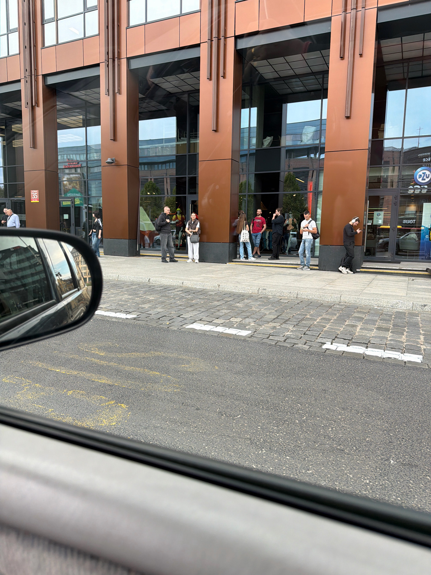 The image captures a street scene in Wrocław, Poland, featuring a modern building with a copper-colored facade and large glass windows. Several individuals are standing on the sidewalk in front of the building, some appearing to be waiting or conversing. One person in the foreground, seen through a car's side mirror, is a man in dark clothing with headphones on, looking at his phone. The sidewalk is paved with cobblestones, and a paved road with yellow markings is visible in the foreground. The reflection in the car's mirror shows buildings and a cloudy sky, suggesting an urban environment. The overall impression is of a typical day in a city, with people going about their business near a commercial or office building.