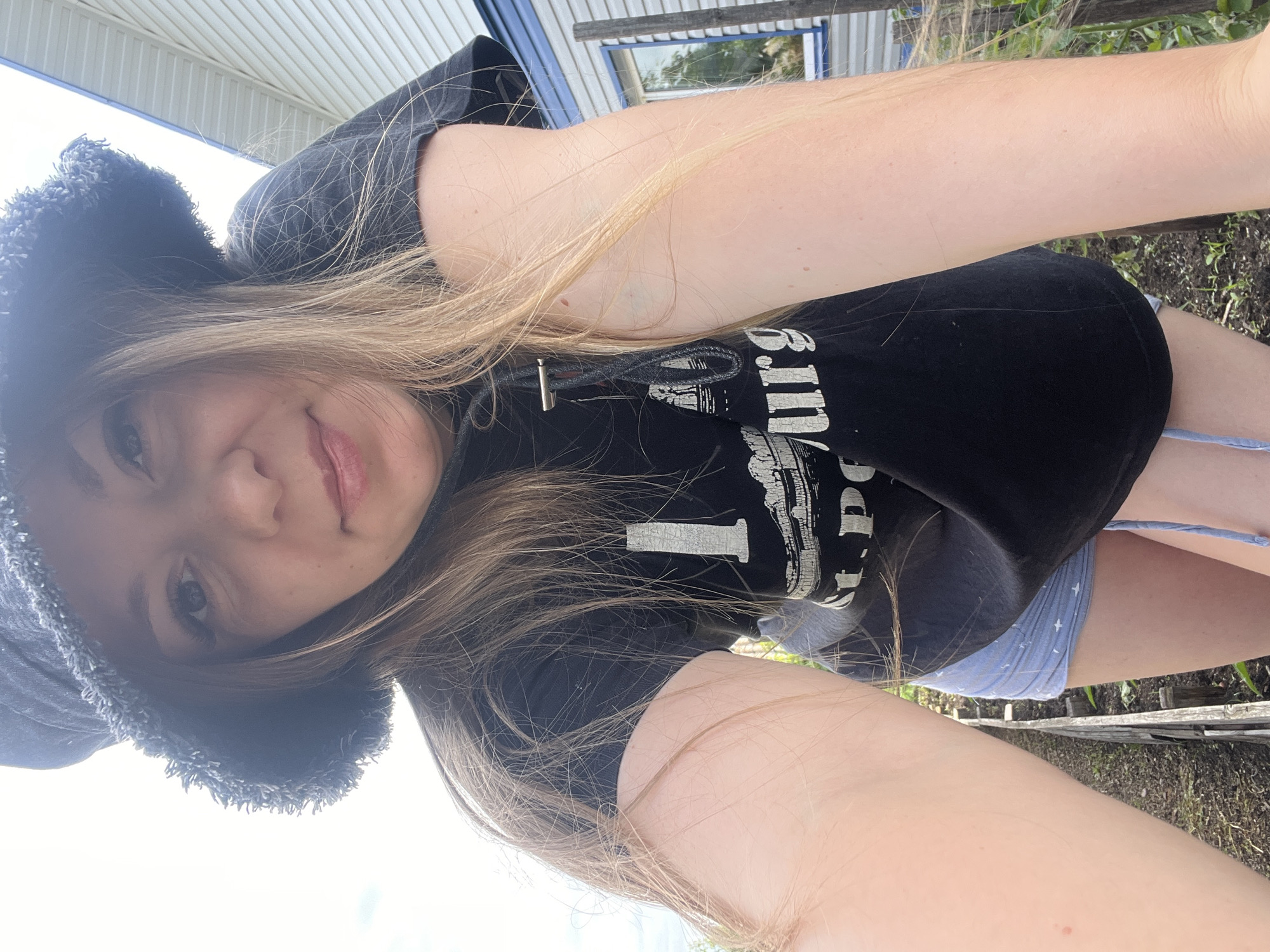 A young Caucasian woman wearing a black bucket hat, a black t-shirt with white lettering that reads "I <3 St. Petersburg," and light blue patterned shorts is pictured outdoors. She is looking at the camera, leaning forward. Her long blonde hair is visible. The background shows a white building with blue trim and a wooden fence. The scene appears to be a garden or backyard in Shadrinsk, Russia.