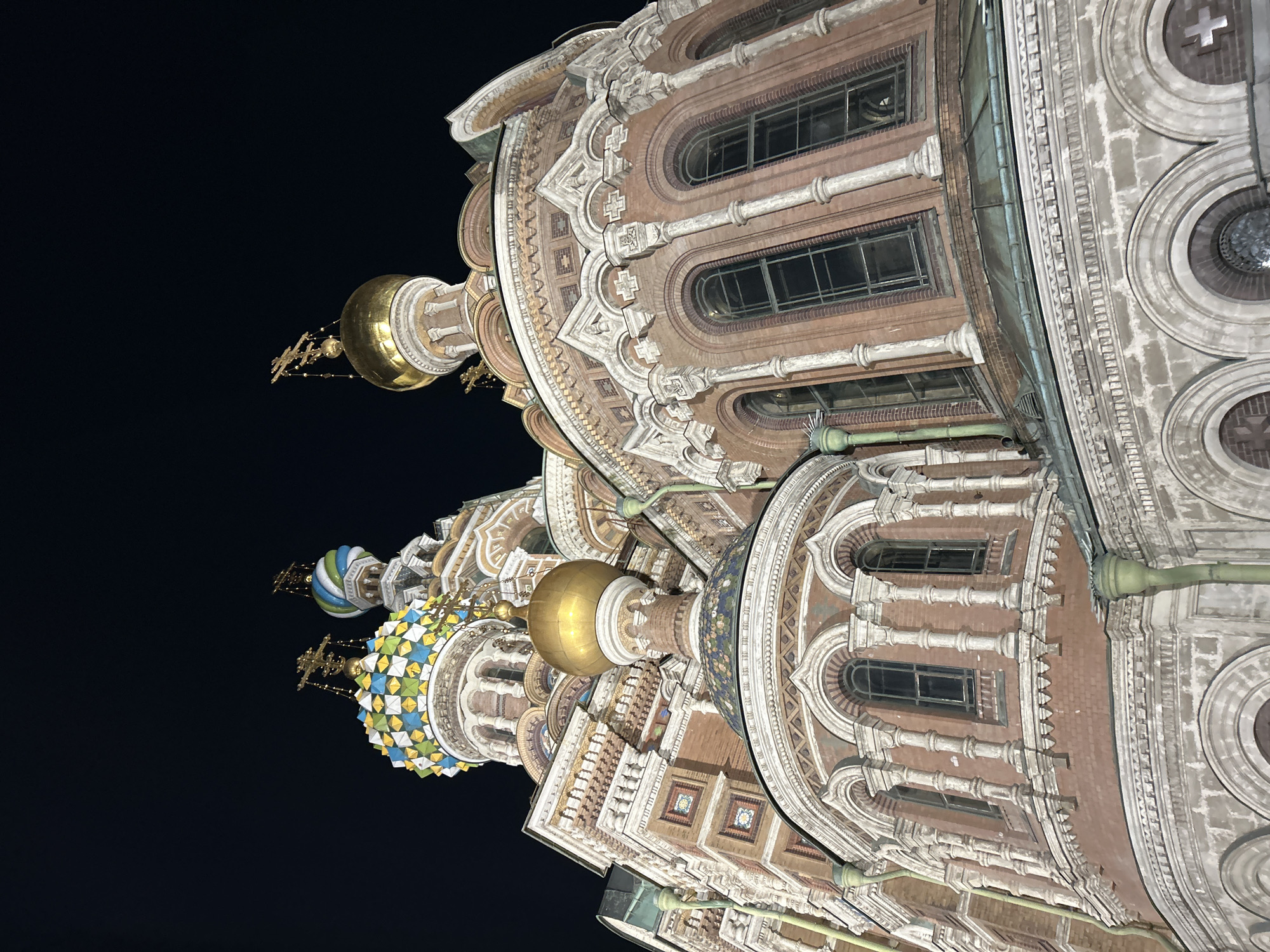 The image depicts a section of the Church of the Savior on Spilled Blood, located in Saint Petersburg, Russia, at night. The architectural details visible include domes with distinct colors and patterns, as well as ornate brickwork, arches, windows, and decorative stone or plaster elements. The sky is dark, emphasizing the illuminated structure.