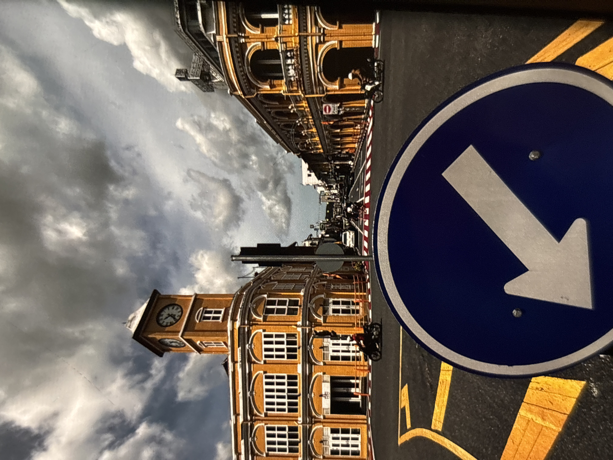 This is a street scene in Chalong, Thailand, featuring a prominent clock tower and colorful Sino-Portuguese architecture. The sky is filled with dramatic, heavy clouds. In the foreground, a blue circular traffic sign with a white arrow pointing downwards and to the left is visible. The road is asphalt with yellow lane markings. A motorcycle with a rider is moving away from the camera on the left side of the road. Further down the street, more buildings with arched windows and balconies line both sides. Another motorcycle is visible on the right side of the street. A white car is also seen in the distance. The overall impression is of a bustling street with historical buildings under a dynamic sky.