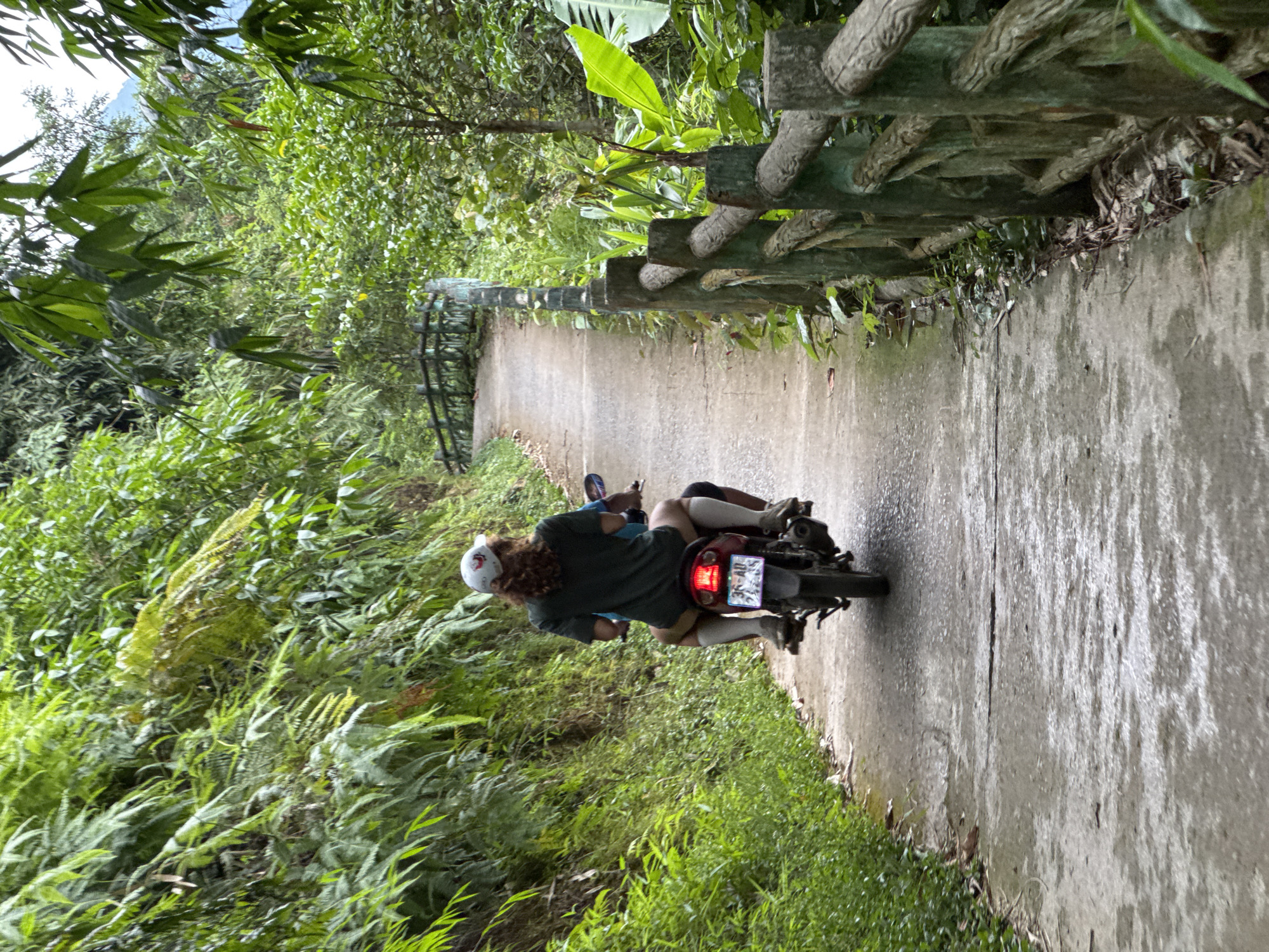 In Pù Luông Commune, Vietnam, a person wearing a dark green t-shirt, white helmet, and white knee-high socks rides on the back of a motorcycle. The motorcycle is red with a license plate that reads "36-AD 242.35". The rider's arms are around the person driving the motorcycle, who is only partially visible. They are on a wet, paved road that curves through a lush, green environment with dense vegetation on both sides. A concrete and wood-like fence runs along the right side of the road, separating it from more foliage and trees. The overall scene depicts a moment of travel through a natural, rural setting.