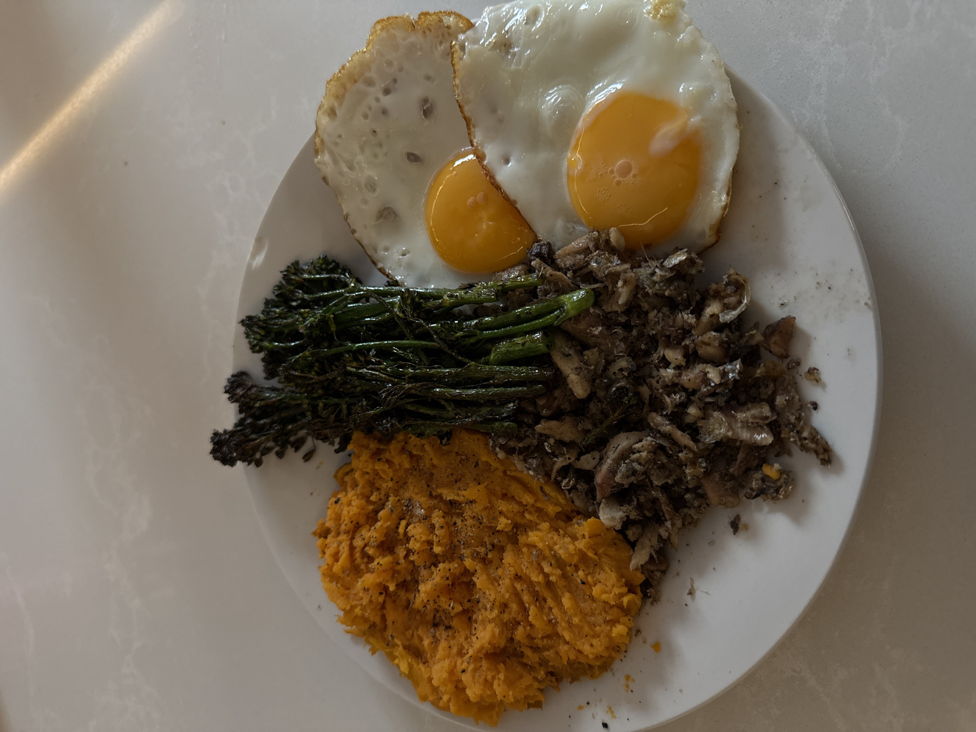 A top-down, close-up shot depicts a white plate arranged with food items, likely a meal. The food comprises two fried eggs with runny yolks, positioned at the top. Adjacent to the eggs is a portion of charred broccolini. Below the broccolini, at the bottom of the plate, sits a mound of mashed sweet potato. To the right of the broccolini and sweet potato is a serving of shredded fish. The plate rests on a white surface, suggesting a kitchen or dining area. The scene does not provide any cues about location in London, United Kingdom, nor does it depict any people, actions, or interactions.