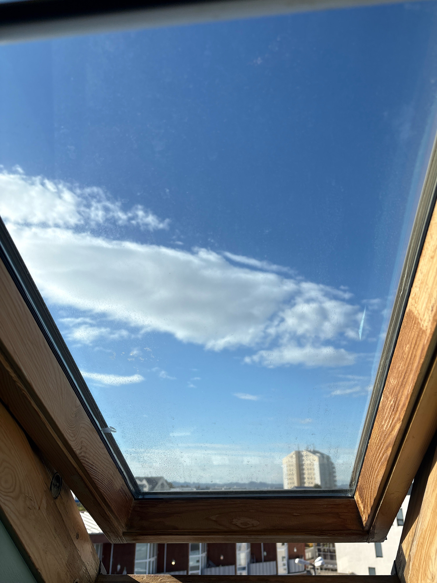 The image shows a view through a slightly dirty skylight, looking out at a bright blue sky with scattered white clouds. In the distance, a multi-story building with a light-colored facade is visible. The skylight frame is made of natural wood. The scene does not contain any people or indicate any specific actions or interactions. The environmental context suggests an urban setting with buildings and a clear day. This scene could be in Reykjavik, Iceland, given the building architecture and sky conditions typical of the region.