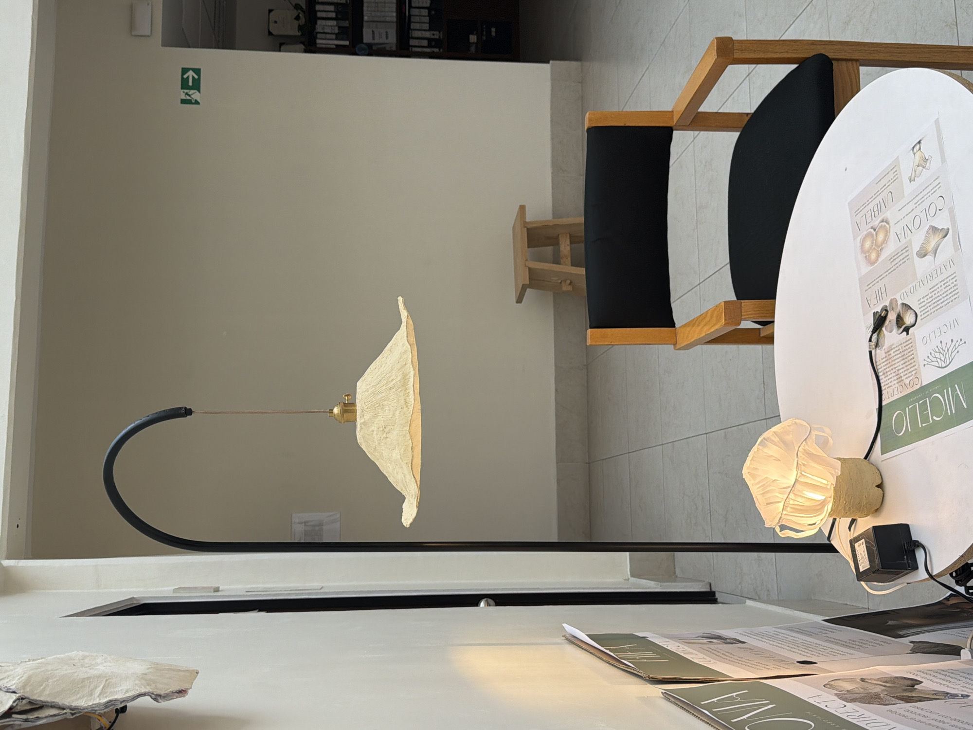 The image shows a dimly lit room, likely indoors, in San Andrés Cholula, Mexico. It features a lamp with a unique, mushroom-shaped shade, positioned above a white table. On the table, there are several documents or leaflets, possibly related to the lamp's design or origin. A black chair with a wooden frame is visible in the background, along with a small wooden stool and a white round table with some leaflets on it. The overall environment suggests a gallery or design studio, with an emphasis on lighting and art. An exit or stair direction sign is visible on the wall.
