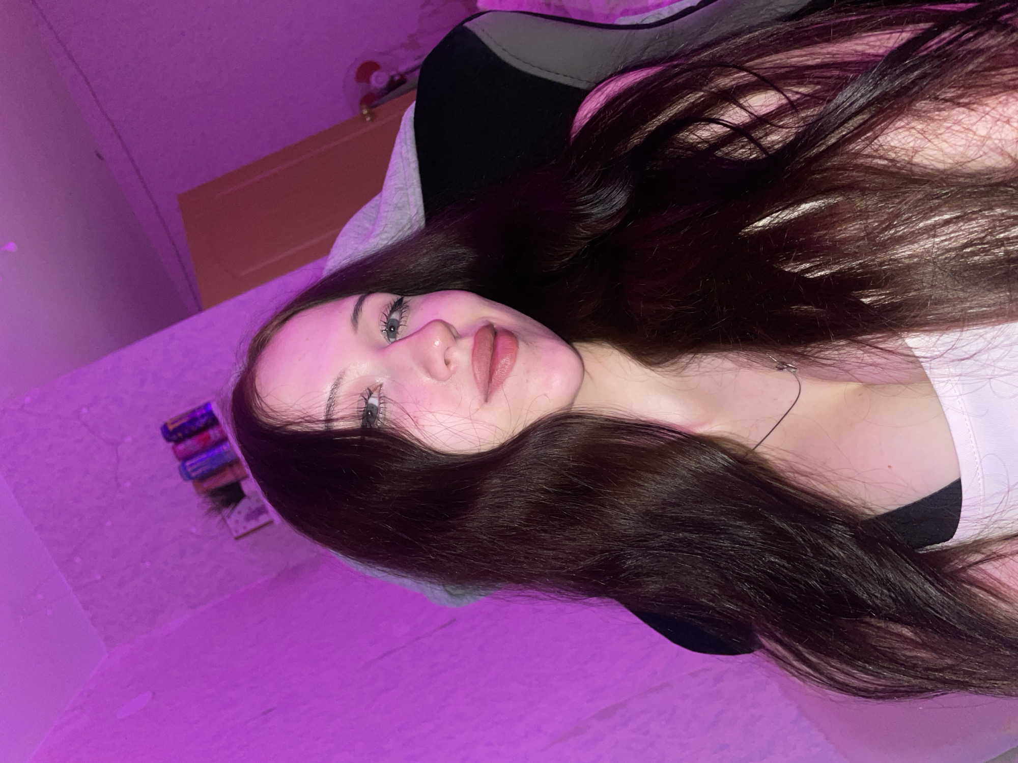 A young woman with long, dark hair and light blue eyes is captured in a close-up selfie. She is wearing a light-colored top and a dark bra. The background is illuminated by pink lighting, creating a vibrant and slightly surreal atmosphere. To the left, a textured wall is visible. On a shelf above her, several cans and a small potted plant are arranged. To the right, a door with a light brown finish is partially visible, along with the back of a dark chair with gray accents. The overall setting appears to be an indoor residential space, possibly a bedroom or gaming area. There is no indication of the city of Votkinsk, Russia, within the image itself.