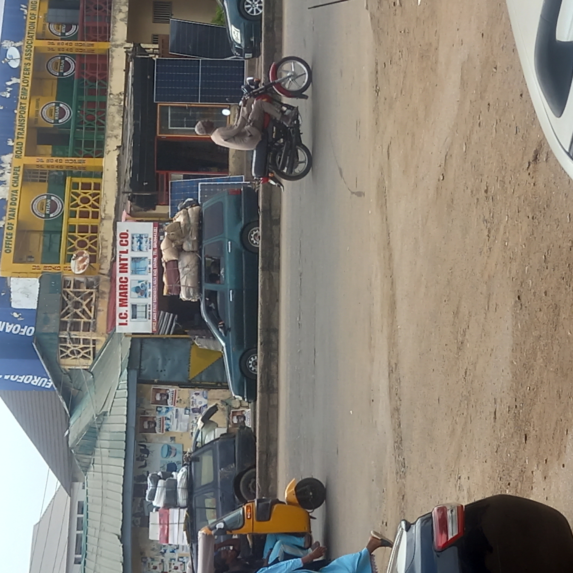 This image captures a bustling daytime street scene in what appears to be an urban or semi-urban area, likely in Sardauna, Nigeria. The sun is bright, suggesting clear weather conditions.
The scene features several vehicles and people. A man, wearing a light-colored top and a cap, is seated on a red and black motorcycle, positioned in the middle of a dirt and asphalt road. Further down the road, a yellow three-wheeled taxi (keke napep) is visible, with a person in blue attire standing or sitting near it. Behind the keke napep, a dark-colored SUV-like vehicle is parked, and another blue car is partially visible in the foreground. A green SUV-like vehicle is parked along the roadside, heavily loaded with bundles tied to its roof rack.
On the left side of the road, several buildings line the street. One prominent building is bright yellow and green, bearing the large blue text "OFFICE OF YAN DOYA CHAPEL ROAD TRANSPORT EMPLOYER'S ASSOCIATION OF NIG". This building also displays several circular logos with the text "RTEAN". Adjacent to it, a large white sign for "I.C. MARC INT'L CO." is clearly visible, with smaller text below it stating "OFFICE LAGOS STREET MUBARAKAT, AMOYE ROAD, KADUNA, TEL: 08039441282". Several solar panels are installed on the roofs or walls of the buildings. Other visible, partial texts include "EUROFO" and "OFOAN", possibly from a "EUROFOAM" sign. The general atmosphere suggests routine daily activities and commerce in a lively street environment.