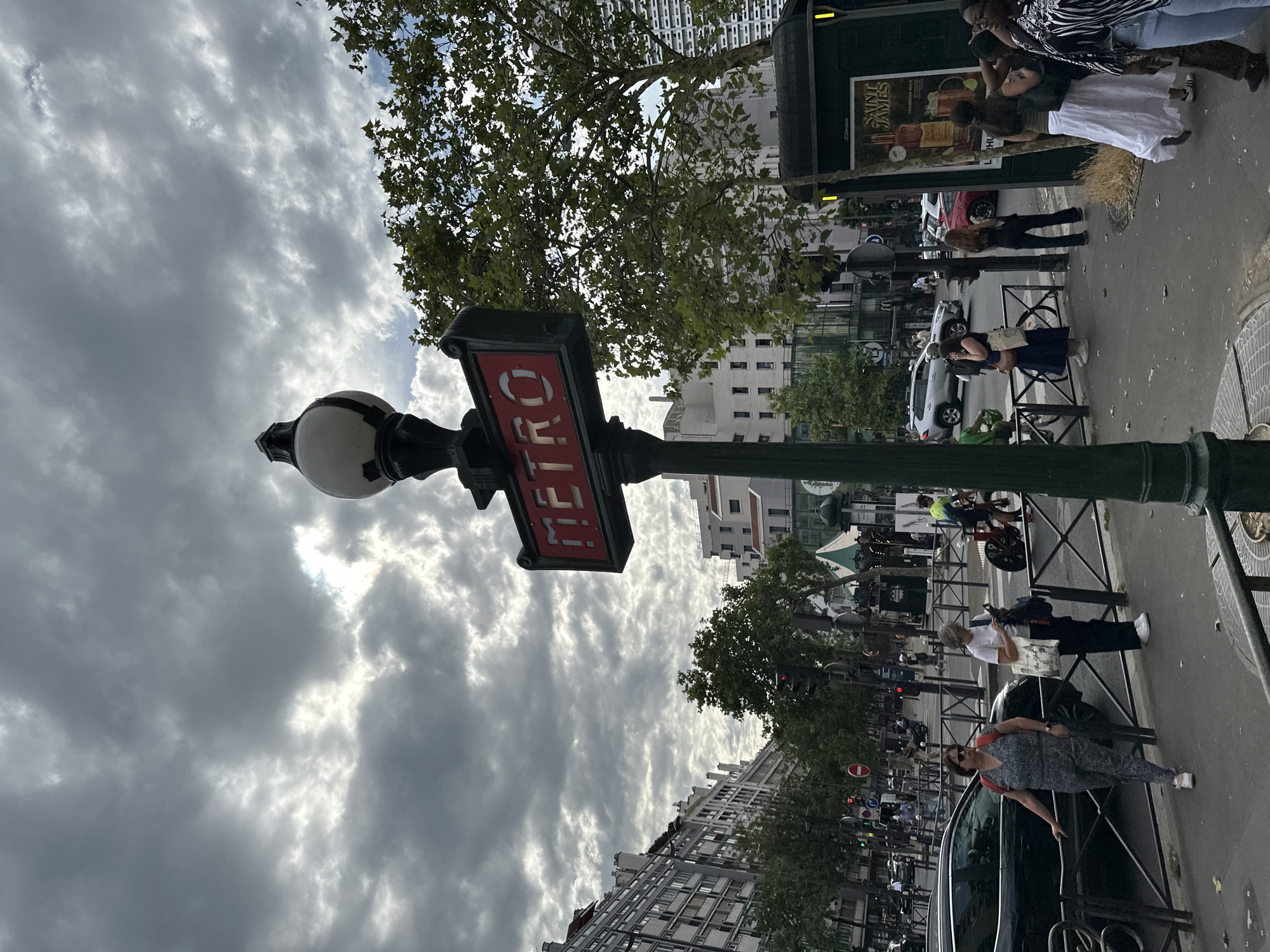 This is a vertical, outdoor photograph of a street scene in Paris, France, under a cloudy sky. The primary subject is a classic green Parisian Metro sign with a red illuminated "METRO" sign. A street lamp with a white globe sits atop the Metro signpost.
Several people are visible in the scene, mostly walking on the sidewalk or near the street. On the right side of the image, a group of people stand near a bus stop or kiosk with advertisements. One person wears a white dress, another a black and white striped top, and others are partially visible. Further down the sidewalk, a woman in a blue dress leans against a railing, looking at her phone. Another woman in a patterned jumpsuit stands near a black car.
In the background, buildings with apartment windows line the street, characteristic of Parisian architecture. Trees with green leaves are scattered along the sidewalk. Cars and traffic lights are also visible, indicating an urban environment.
The time of day appears to be daylight, but the heavy cloud cover creates a somewhat dim and overcast atmosphere. The overall mood is that of a typical, busy day in a city.
Visible text includes "METRO" on the sign and portions of advertisements on the kiosk, which seem to include text like "SAINT JAMES". Traffic signs with circles and bars are also present.
