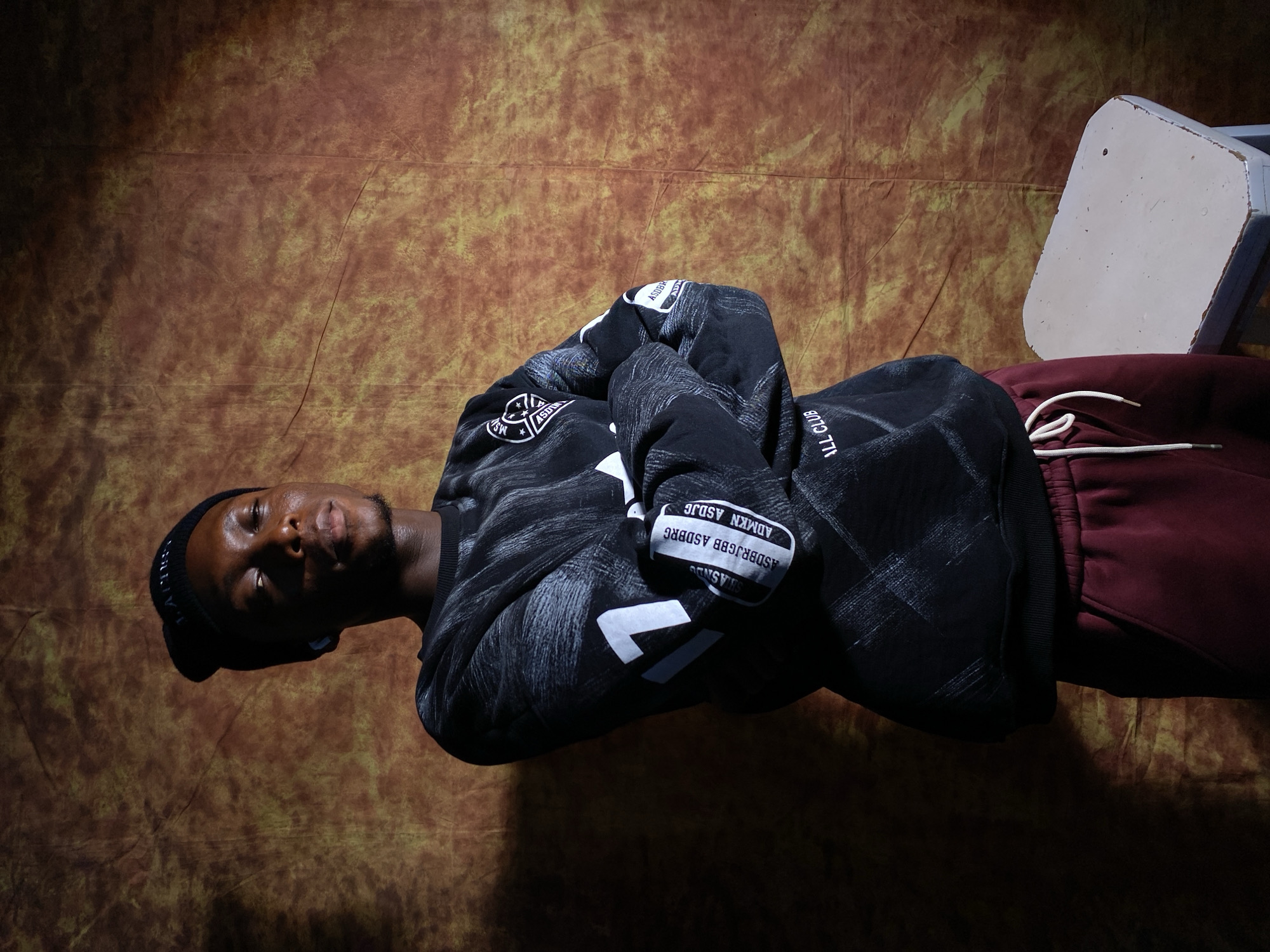 A young Black man stands with his arms crossed in front of a textured, orange-brown backdrop. He is wearing a black beanie, a black sweatshirt with a white graphic pattern and text, and maroon sweatpants. He appears to be wearing earbuds. To his right, a small, white stool is visible. The lighting suggests an indoor setting, possibly a studio, given the backdrop. The location is not specified in the image.
