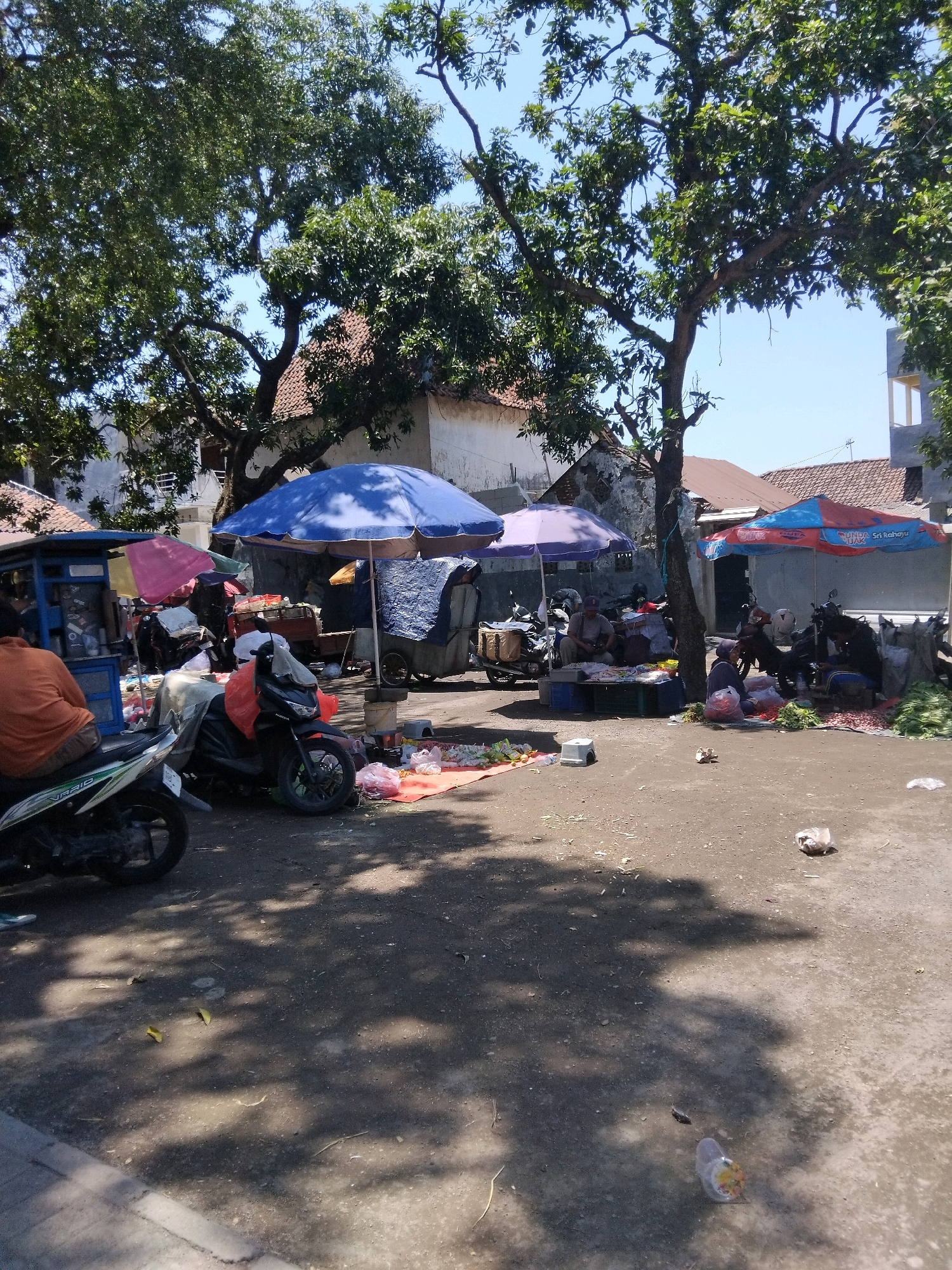 This is a scene from the Kepuh Kiriman market in Indonesia, captured under a clear blue sky with abundant sunlight casting shadows. The foreground is dominated by a dirt and gravel ground, with a paved walkway on the left edge. Scattered across the ground are dry leaves and plastic waste.
The midground of the image reveals a bustling market setting. Several motorbikes are parked, some covered. Tents and umbrellas, in blue, pink, and red/blue patterns, provide shade for various stalls. Under these shelters, vendors are present, some seated on the ground, others behind their merchandise. The stalls display a variety of goods, appearing to be fresh produce such as onions, vegetables, and possibly other food items. A person in an orange shirt is seated on a motorbike, facing away from the camera. In the background, buildings with terracotta tiled roofs are visible, interspersed with lush green trees. The overall impression is one of a typical outdoor market scene in Indonesia.