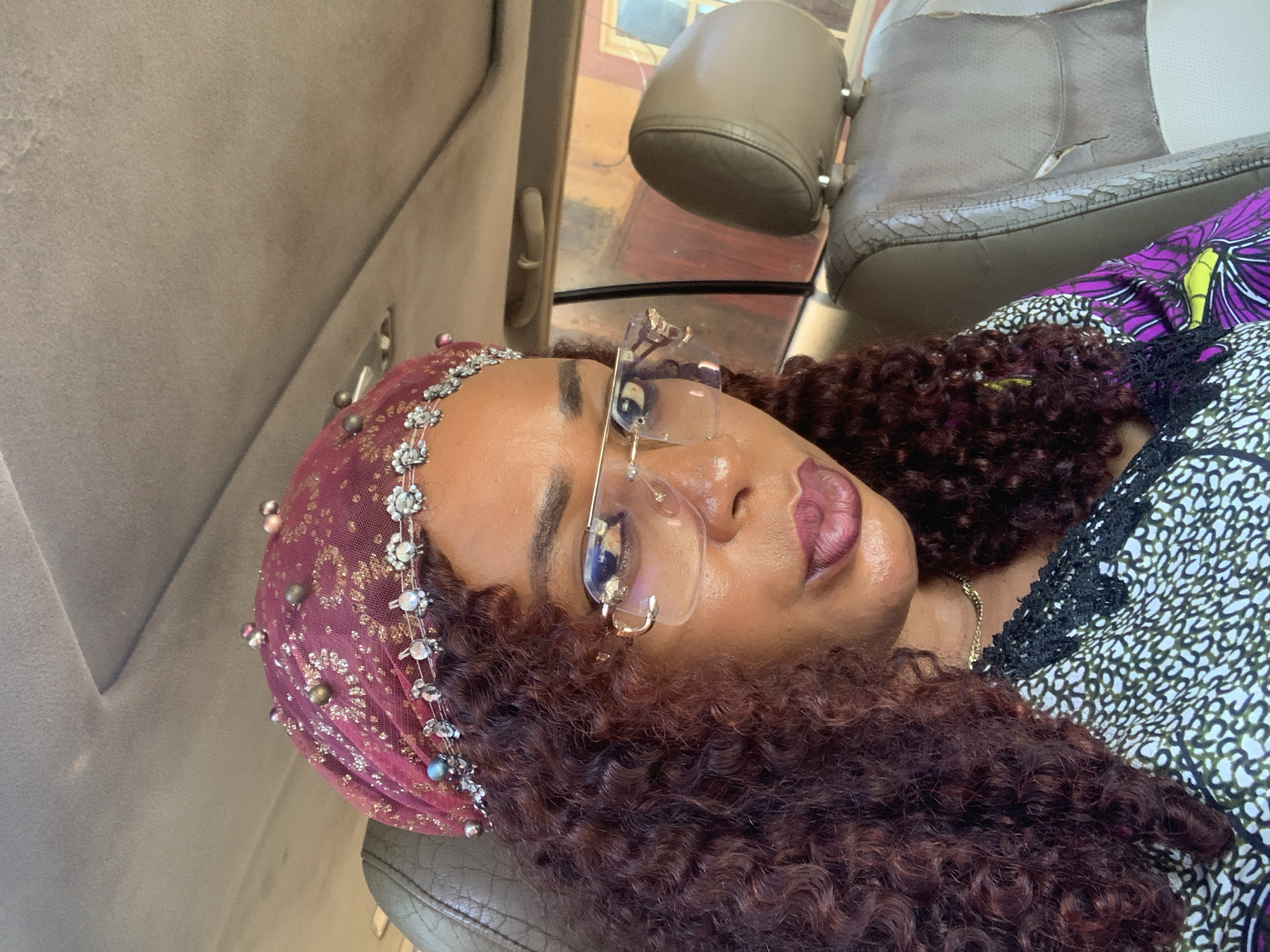 A forensic examination of the image reveals a detailed portrait of a female subject seated within a vehicle. The scene is situated in what is identified as Sabon Tasha, Nigeria.
The subject, a woman with medium-toned skin, occupies the foreground. She possesses voluminous, curly hair in a dark reddish-brown hue, which frames her face. Her head is adorned with a maroon or burgundy headwrap, appearing to be crafted from a sheer, netted fabric embellished with gold-toned glitter or small sequins. This headwrap is further decorated with various small, spherical beads, some appearing metallic and others pearlescent, intermittently placed across its surface. Along the front edge of the headwrap, a decorative band of silver-toned elements, possibly rhinestones or small crystals, is visible.
She wears rimless eyeglasses with gold-toned metal temples, reflecting ambient light and indistinct reflections. Her lips are painted with a dark plum or berry-colored lipstick and are slightly pursed. A well-groomed eyebrow is visible above the left eye. Around her neck, a slender gold-colored chain necklace is present.
Her attire includes a top with a multi-patterned design. The main body of the garment features a white background with an abstract pattern of black and olive-green shapes, and a black lace or trim detail at the neckline. The visible sleeve displays a vibrant pattern incorporating purple and yellow/lime green butterfly or floral motifs.
The immediate environment is the interior of a car. The overhead headliner is light gray or beige and exhibits signs of wear, including minor discoloration and texture variations. The car seats, visible as a driver's seat portion and the full passenger seat, are upholstered in light brown or tan leather or leatherette. Both seats show considerable wear, characterized by creasing and prominent cracking, particularly on the passenger seat's backrest and headrest. A light-colored grab handle is visible above the passenger side window.
Through the passenger side window, an external, blurred view suggests an outdoor setting, likely urban or semi-urban, consistent with a city environment. Vague reddish-brown and lighter-toned structures, possibly parts of buildings or walls, are discernible. The overall lighting suggests daylight.
