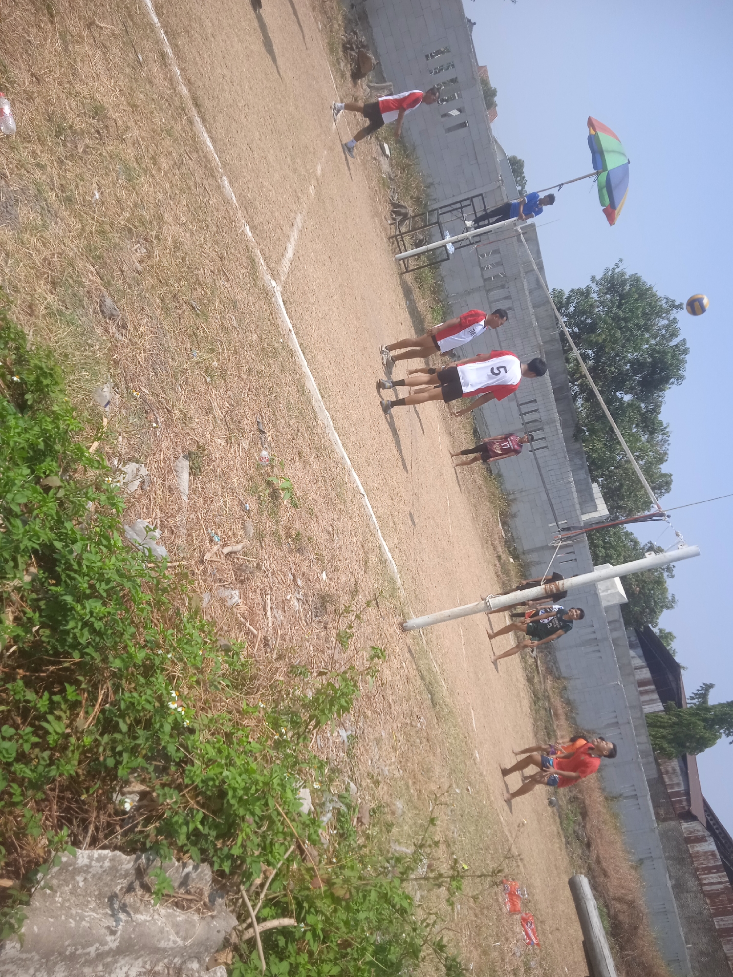 The image depicts an outdoor volleyball game on a dirt court in Klodran, Indonesia. Several individuals are present, actively participating in or observing the game. The court is marked with white lines. A volleyball net is set up across the court, with a volleyball visible in mid-air. A colorful umbrella is mounted on a pole, providing shade. The surroundings include dry grass, sparse vegetation, concrete walls, and trees. The lighting suggests it is daytime with clear skies.