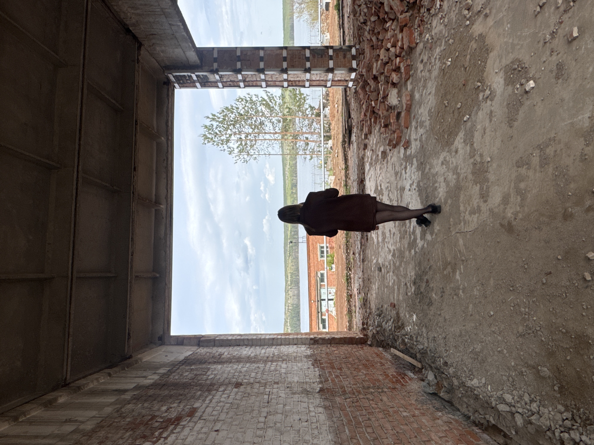 The image depicts a person standing in a ruined, abandoned building in the city of Chaikovsky, Russia. The building appears to be constructed of concrete and brick, with crumbling walls and debris scattered on the floor. The person is wearing a dark-colored coat and tights. Through the window, a view of trees, water and building can be seen. The overall impression is one of decay and desolation.