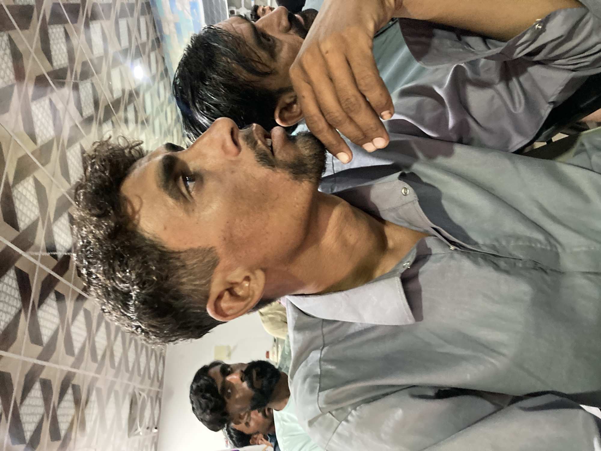 The image shows a group of people, with a man in a gray shirt prominently displayed in the foreground. Another man is partially visible behind him, with a hand resting on his shirt. Additional people can be seen in the background. The image is taken indoors, with a patterned wall in the background. The city and specific location are unknown.