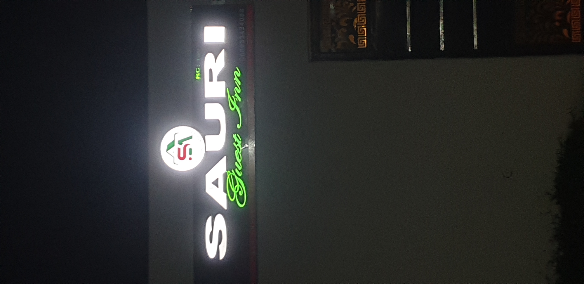 The image shows a nighttime view of a lit-up sign for "SAURI Guest Inn" in Narayi, Nigeria. The sign features the words "SAURI" in large, white, illuminated lettering and "Guest Inn" in green, cursive, illuminated lettering below it. Above the "SAURI" text, there is a circular logo with a stylized house shape and the letters "S" and "A" integrated into it. To the right of the logo, the letters "RC" are visible in smaller, illuminated text, followed by a phone number that appears to be "00933434088". The sign is mounted on a dark surface. To the right of the sign, a dark, vertical structure with horizontal, illuminated bands and a decorative lower section with a Greek key pattern is partially visible. The background is dark, suggesting it is nighttime. There are no people or animals visible in the image.