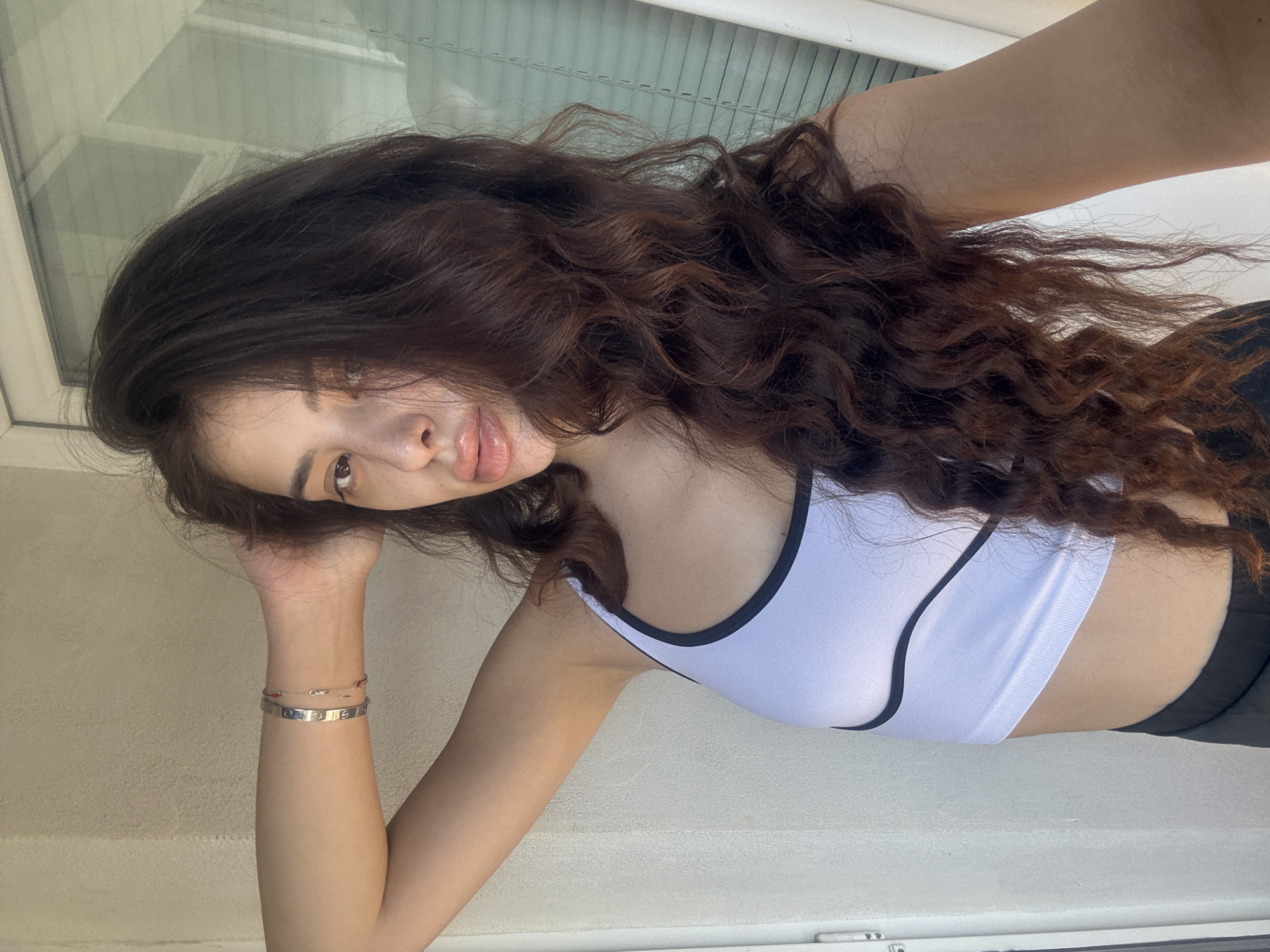 The image is a selfie of a young woman taken outdoors during the day. She has long, wavy, dark brown hair and is wearing a white crop top with black trim and a black bottom. A silver bracelet is visible on her left wrist. She is standing against a light-colored textured wall, with a window or door frame visible to her right. There is no indication of the city of Plovdiv, Bulgaria, in the image.
