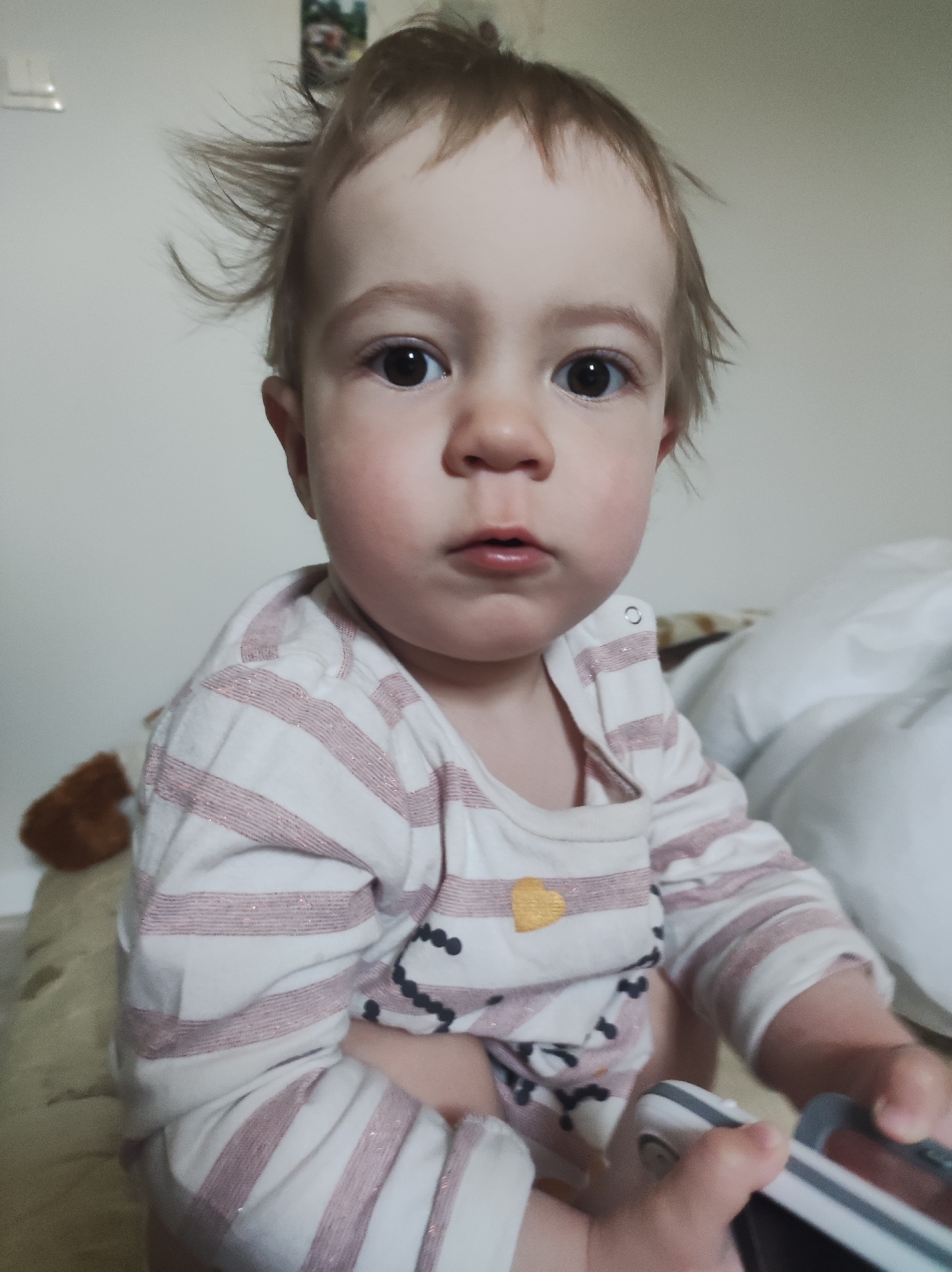 A baby with light brown hair is sitting upright on a bed. The baby is wearing a white long-sleeved shirt with pink stripes and a small yellow heart on the chest. The hair is slightly disheveled. The baby is holding a white and grey object in their hands. A brown teddy bear is visible in the background, along with white pillows. The background wall is white and there is a small picture hanging on the wall. The location is a bedroom. City and specific location are unknown.